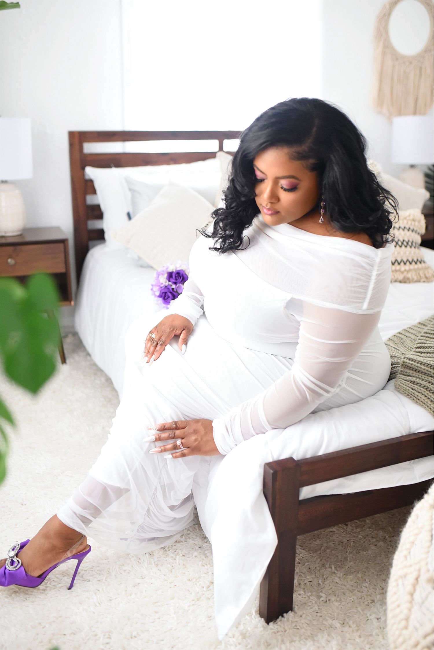 Pregnant woman in a white off-shoulder mesh gown holding purple flowers while seated on a bed.