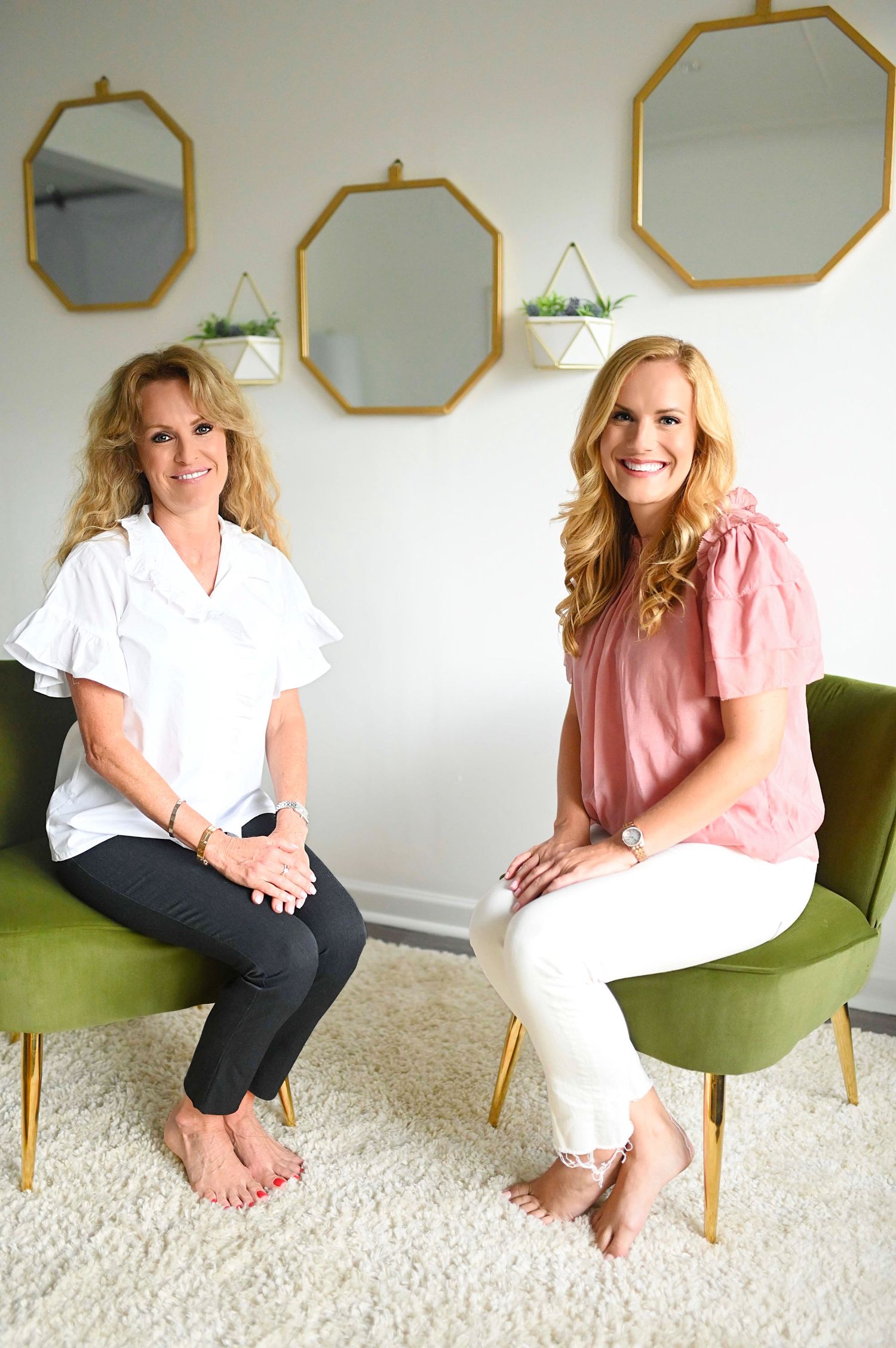Two smiling women seated in green velvet chairs in a bright modern room with gold mirrors.