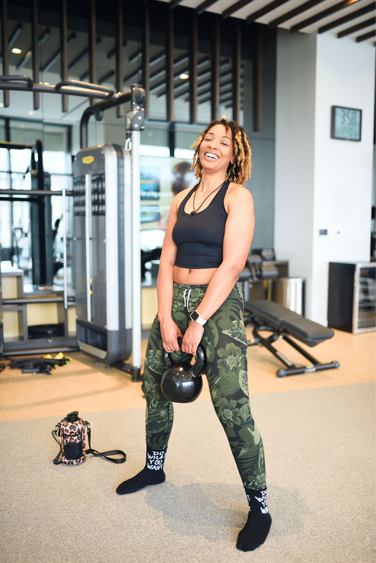 Athletic woman laughing while holding a kettlebell in a modern gym wearing camo leggings and black crop top.