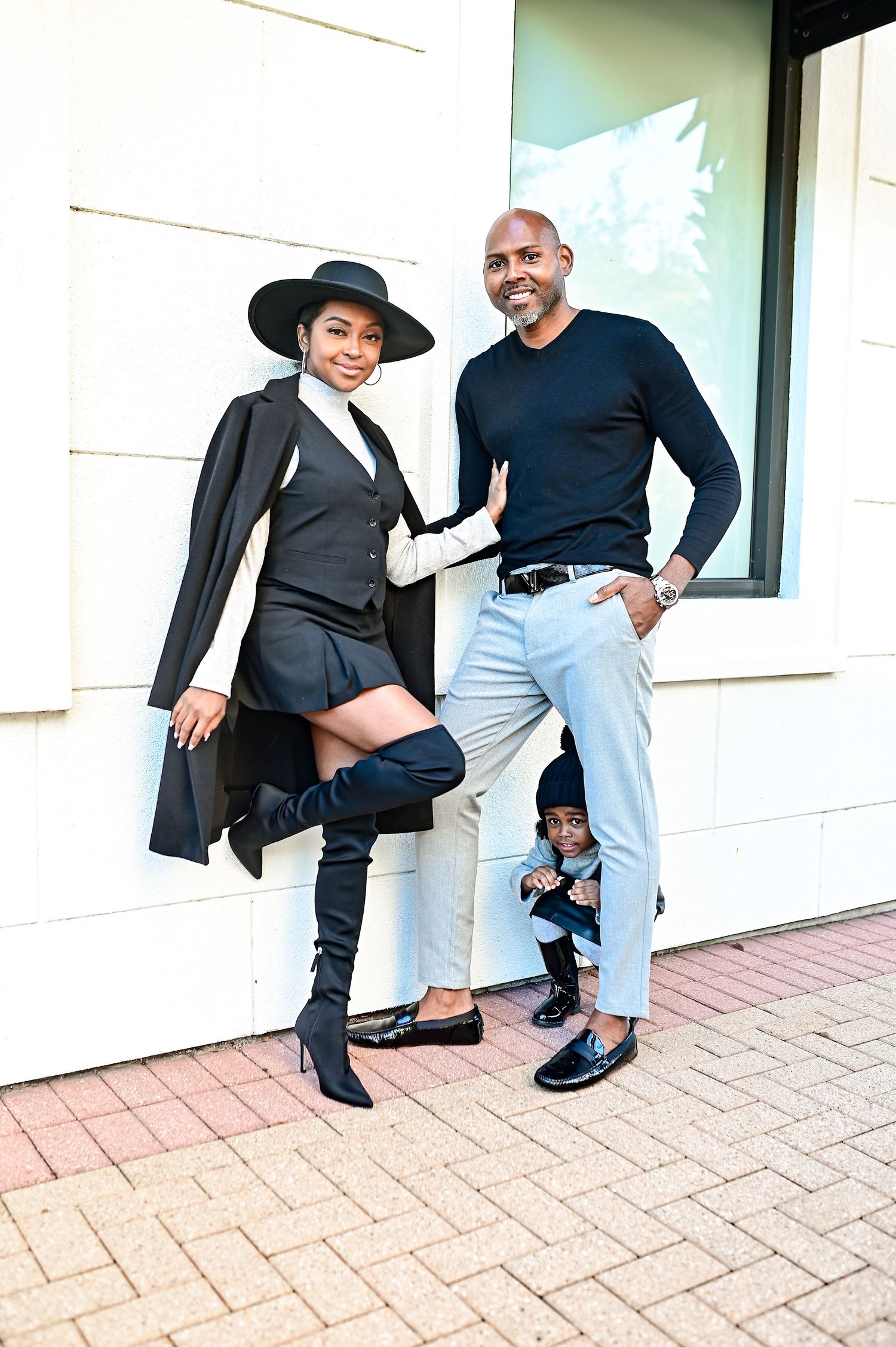 Fashionable couple with a small child posing together outdoors, woman wearing black thigh-high boots and wide-brim hat.