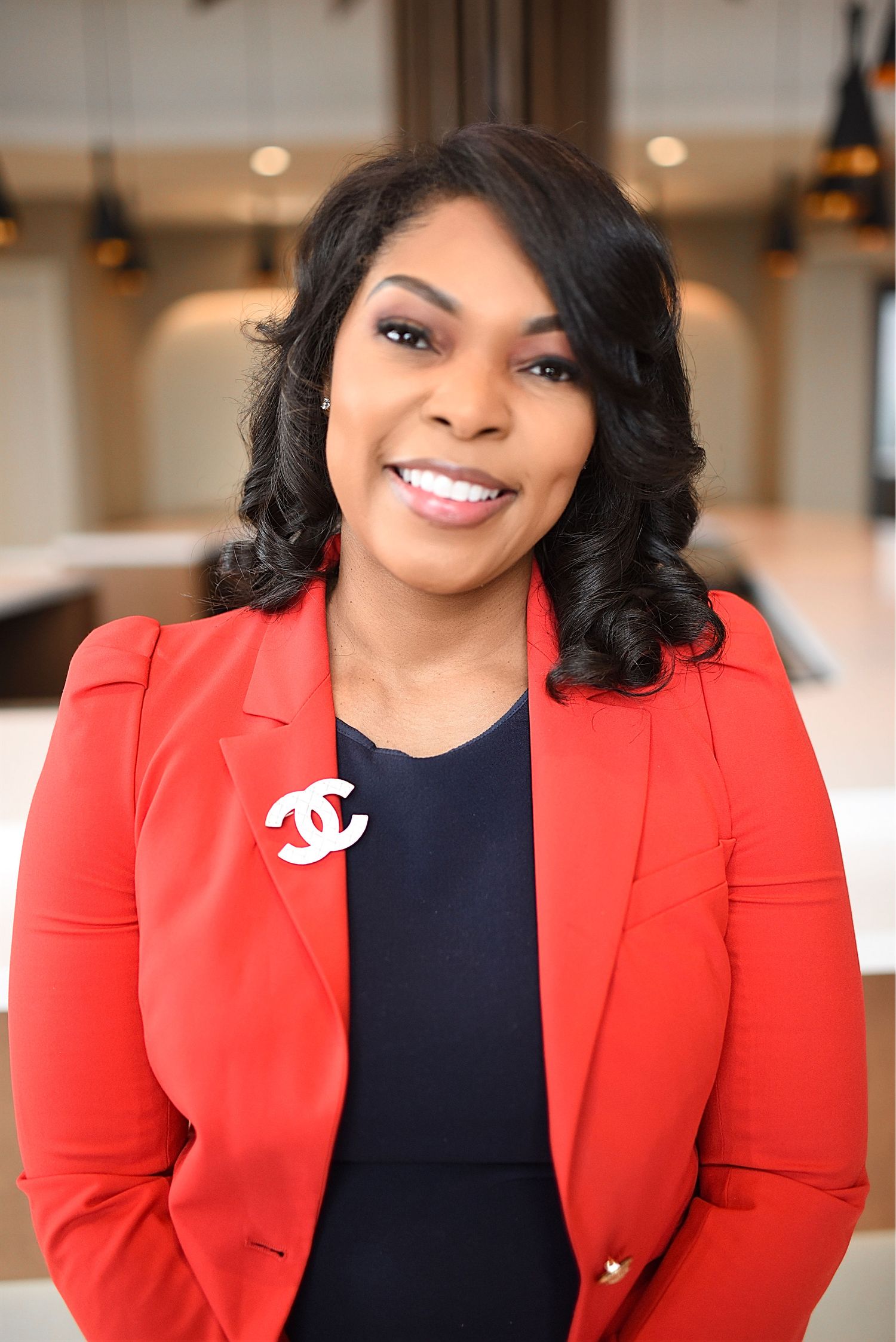 Professional woman in red blazer with Chanel brooch and navy dress smiling in modern interior setting.