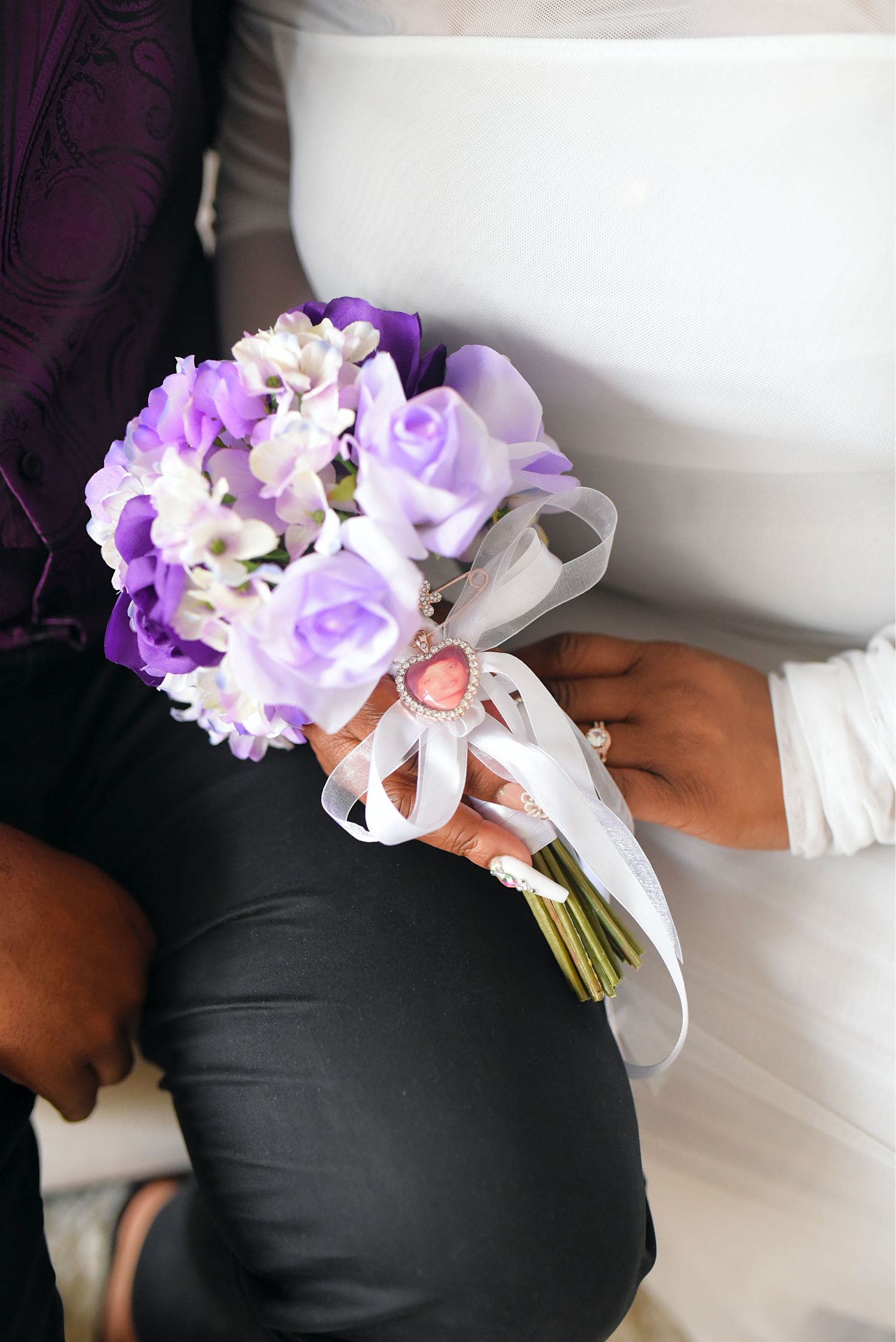 Close-up of hands holding a purple and white rose bridal bouquet with ribbon and pink heart charm.