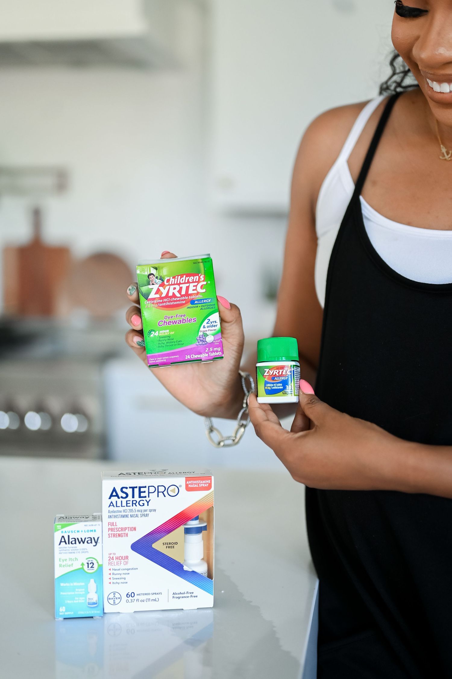 Woman in black dress holding Children's Zyrtec and Zyrtec allergy products in a kitchen.