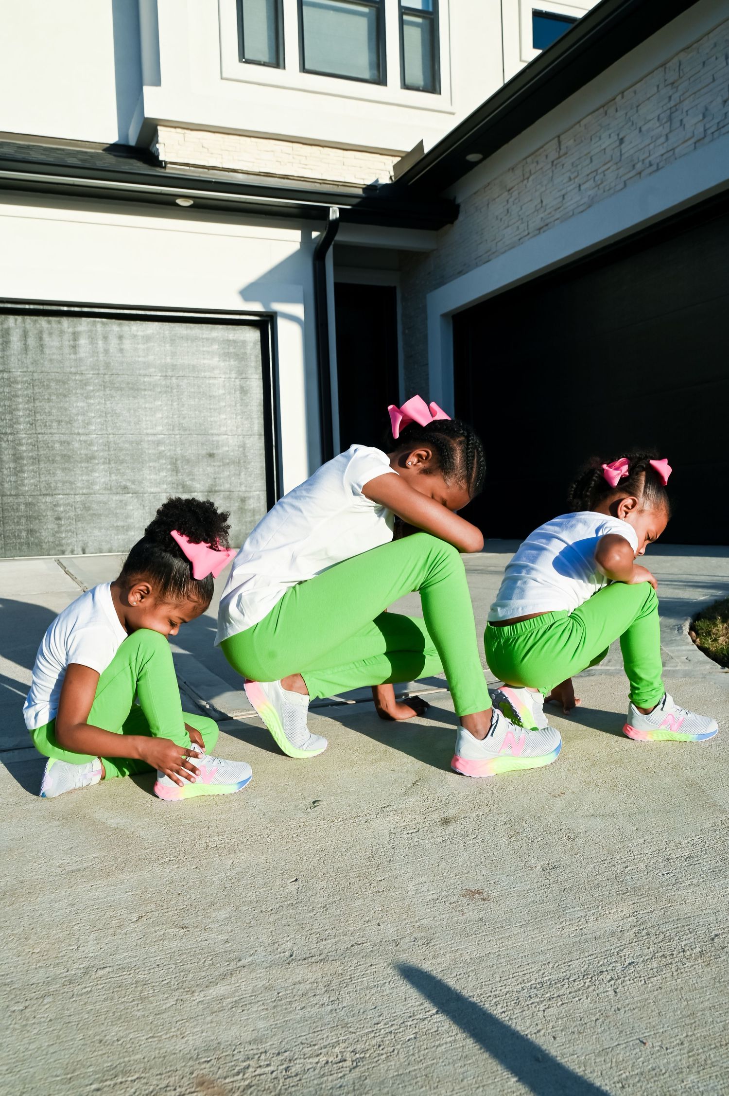 Three young girls in green leggings and pink bows tying colorful sneakers on a driveway.