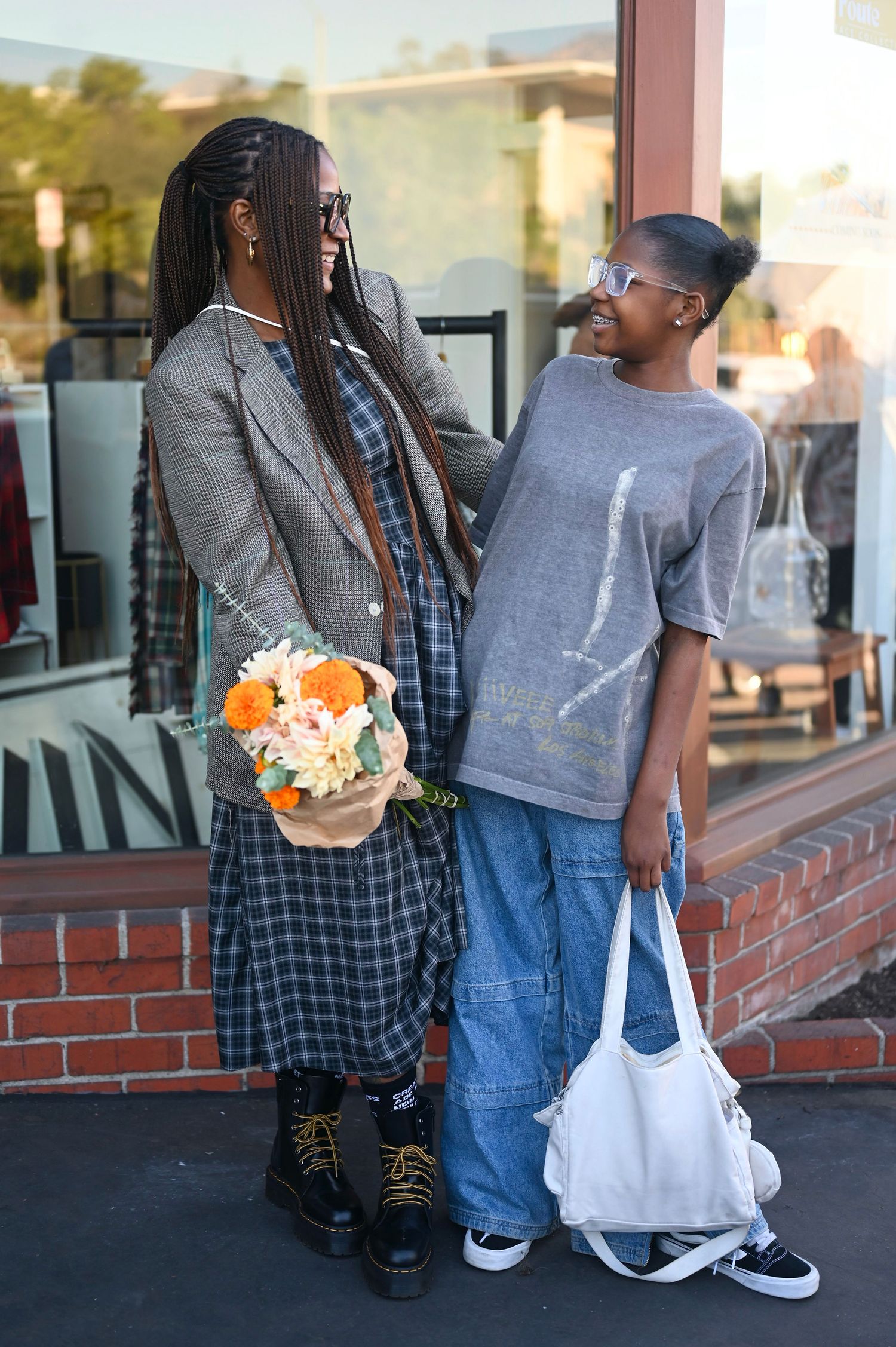 Two stylish young women smiling at each other outside a shop, one holding a bouquet of orange flowers.