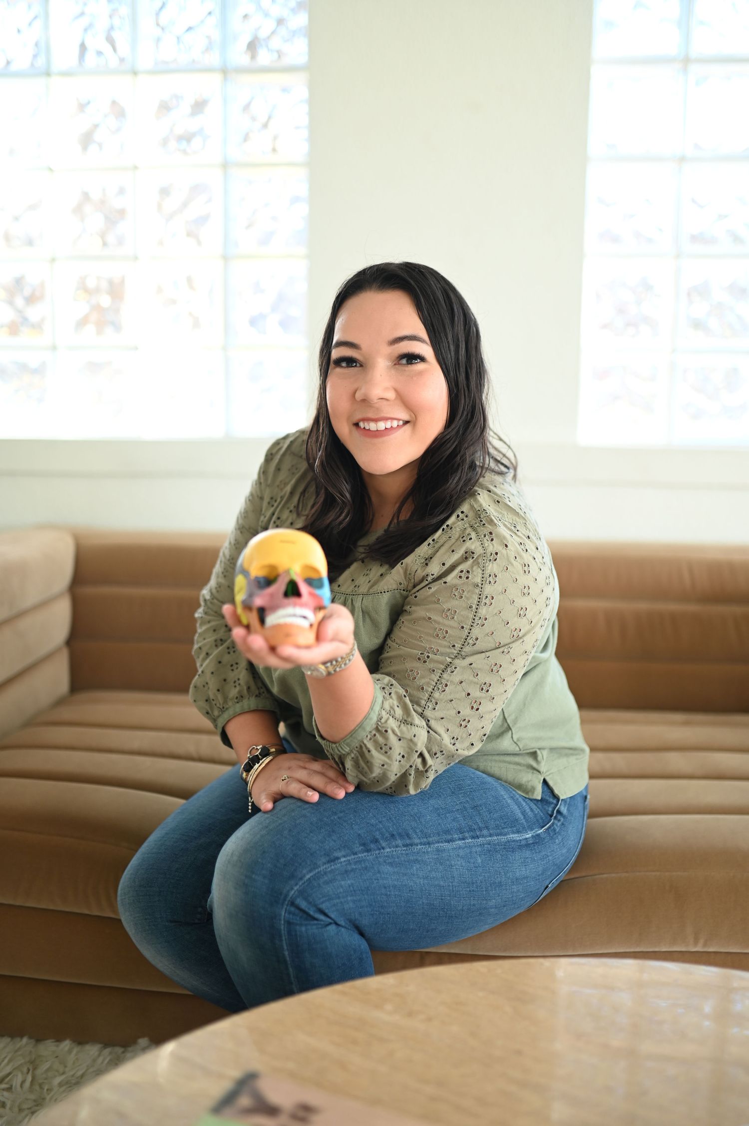 Smiling woman in green sweater and jeans sitting on couch holding a colorful anatomical skull model.