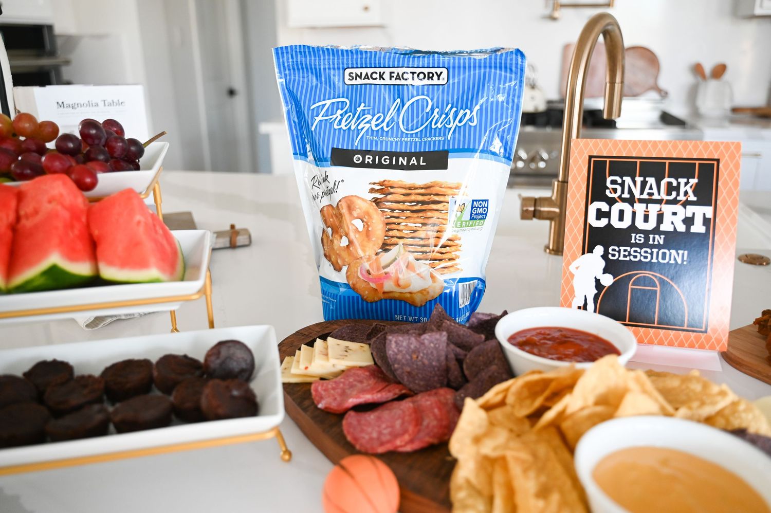 Snack Factory Pretzel Crisps bag displayed with charcuterie board of meats, dips, and fresh fruit.