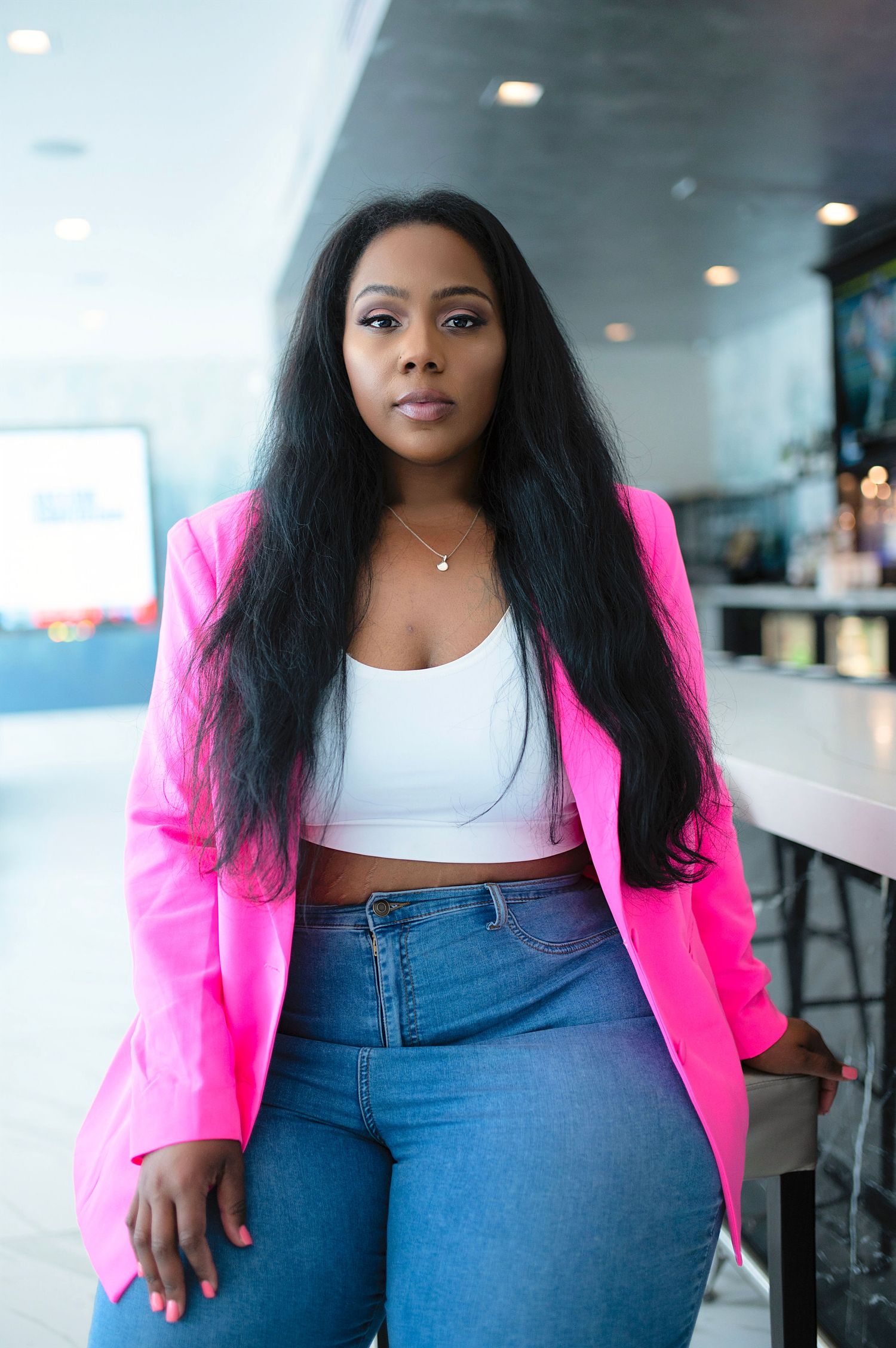 Confident woman in hot pink blazer, white crop top, and jeans posing in modern interior.