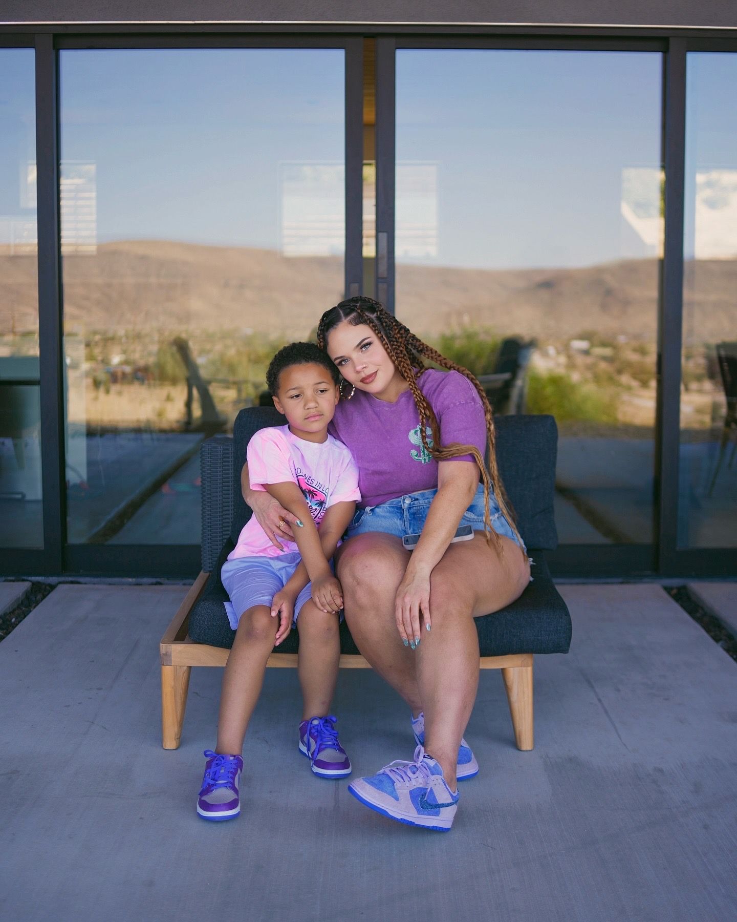 Woman and child in matching purple and pink outfits with blue sneakers sitting on chair with desert mountain backdrop.