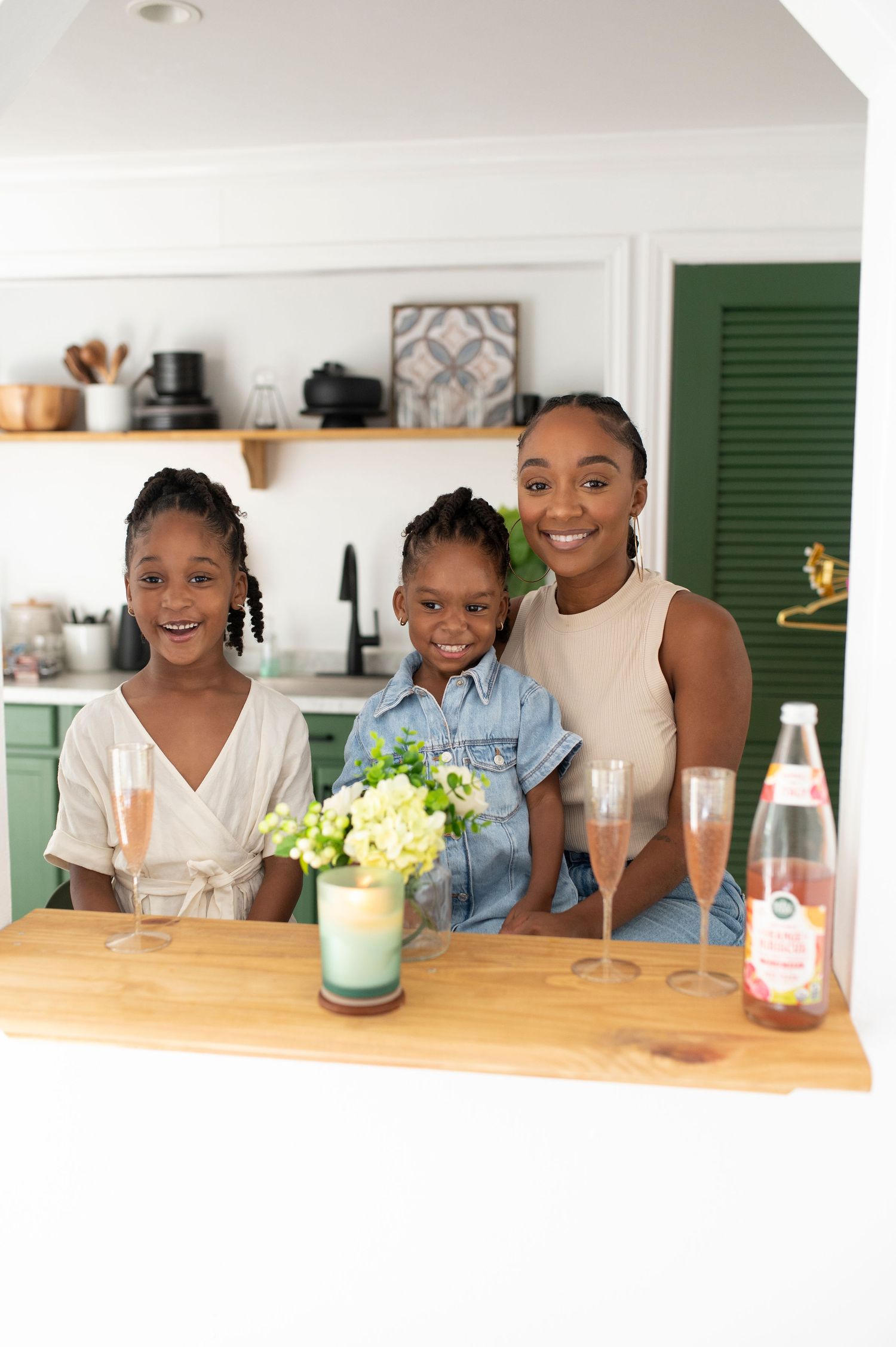 A Black mother and two daughters smile together at a kitchen counter with drinks.