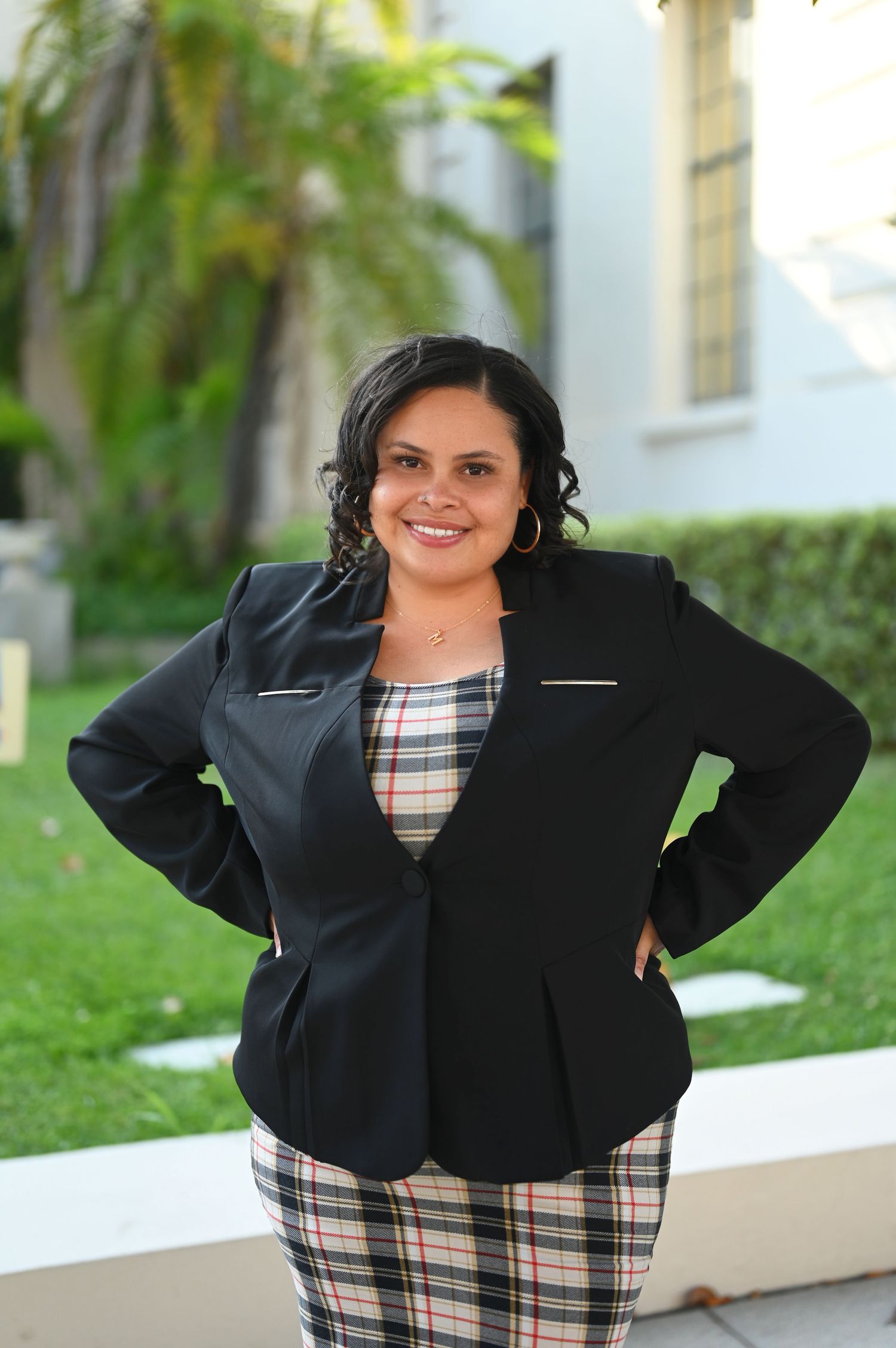 Smiling woman in black blazer and plaid outfit posing with hands on hips in front of green hedges.