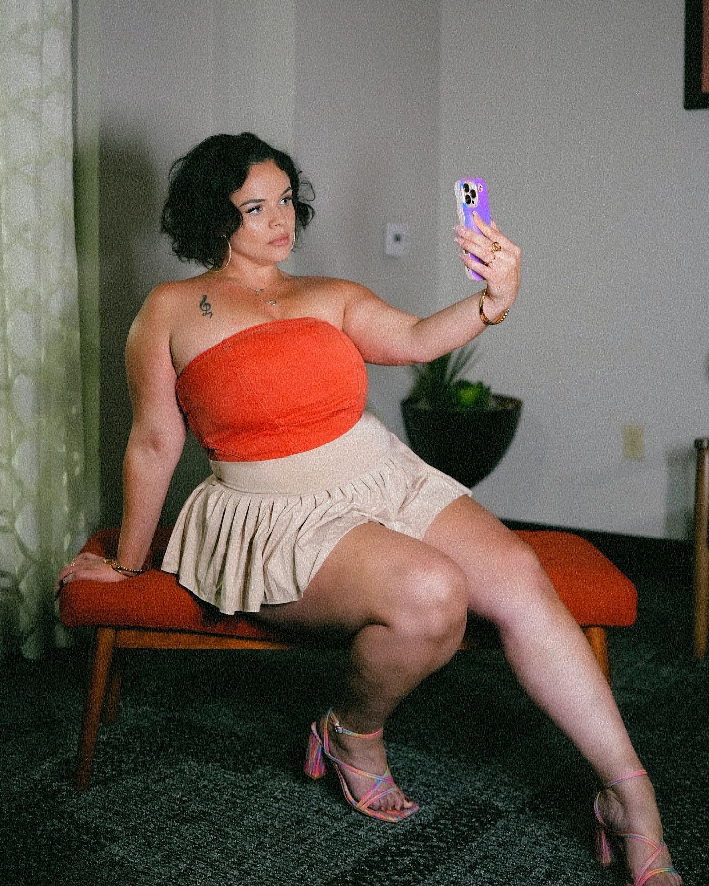 Woman in orange tube top and beige mini skirt takes selfie while seated indoors.