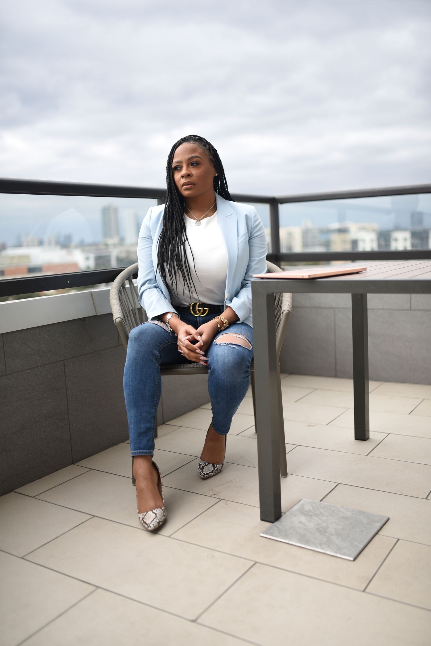 Confident woman in light blue blazer and ripped jeans sitting on rooftop terrace with city skyline backdrop.