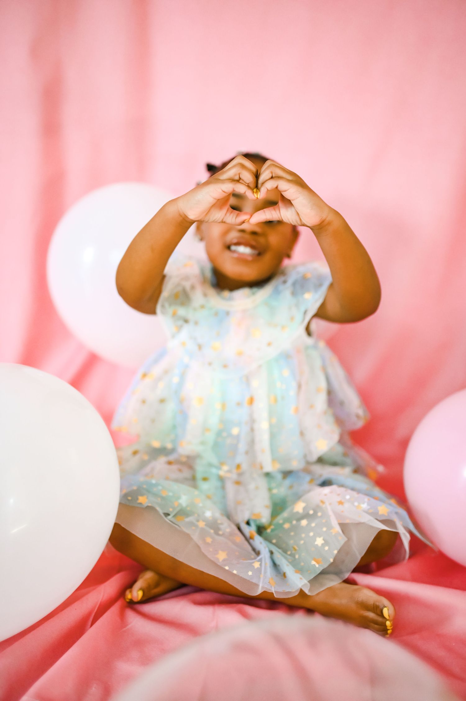 A Black toddler girl makes a heart shape with her hands among white balloons.