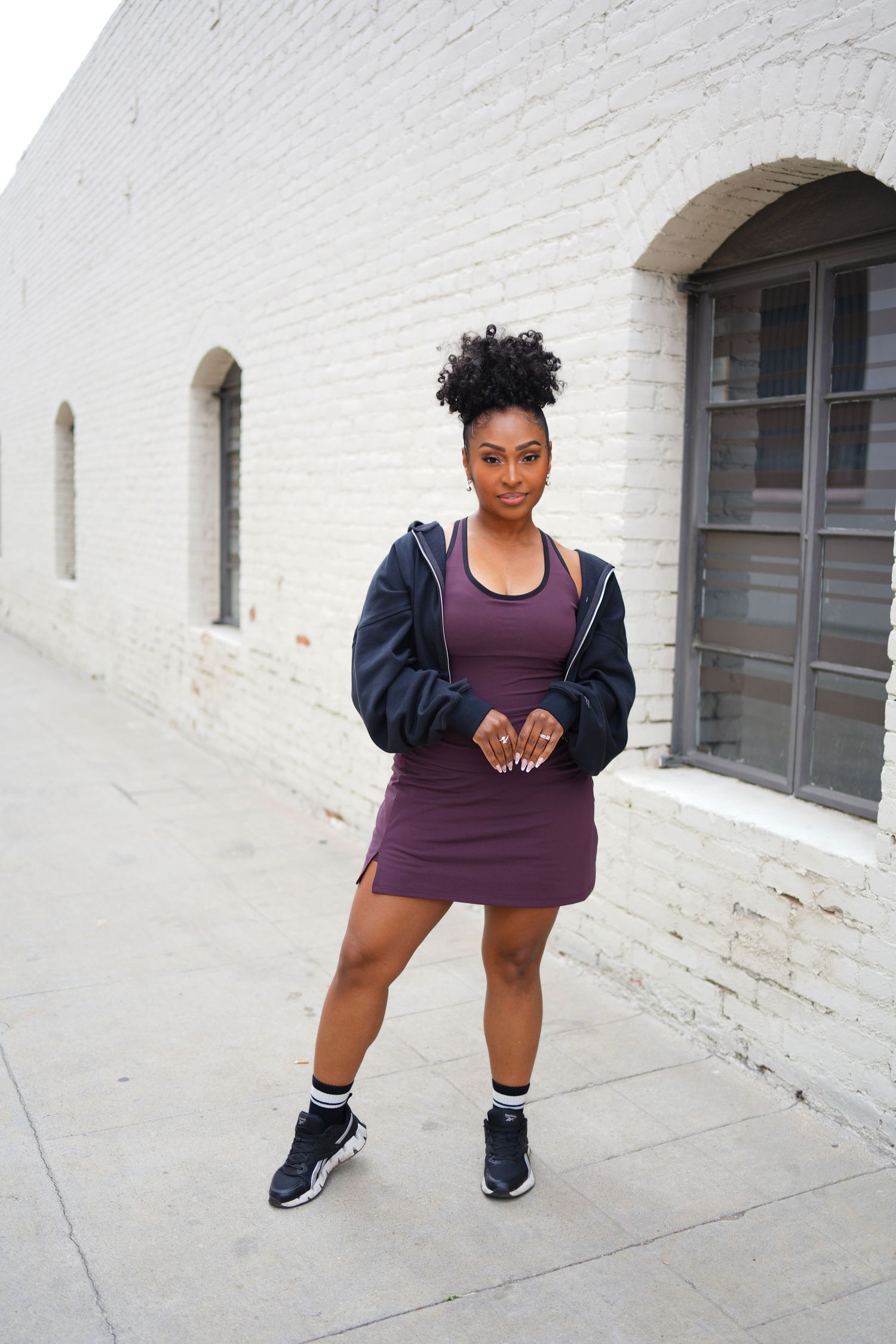 Stylish Black woman in purple dress and navy jacket poses confidently outside white brick building.