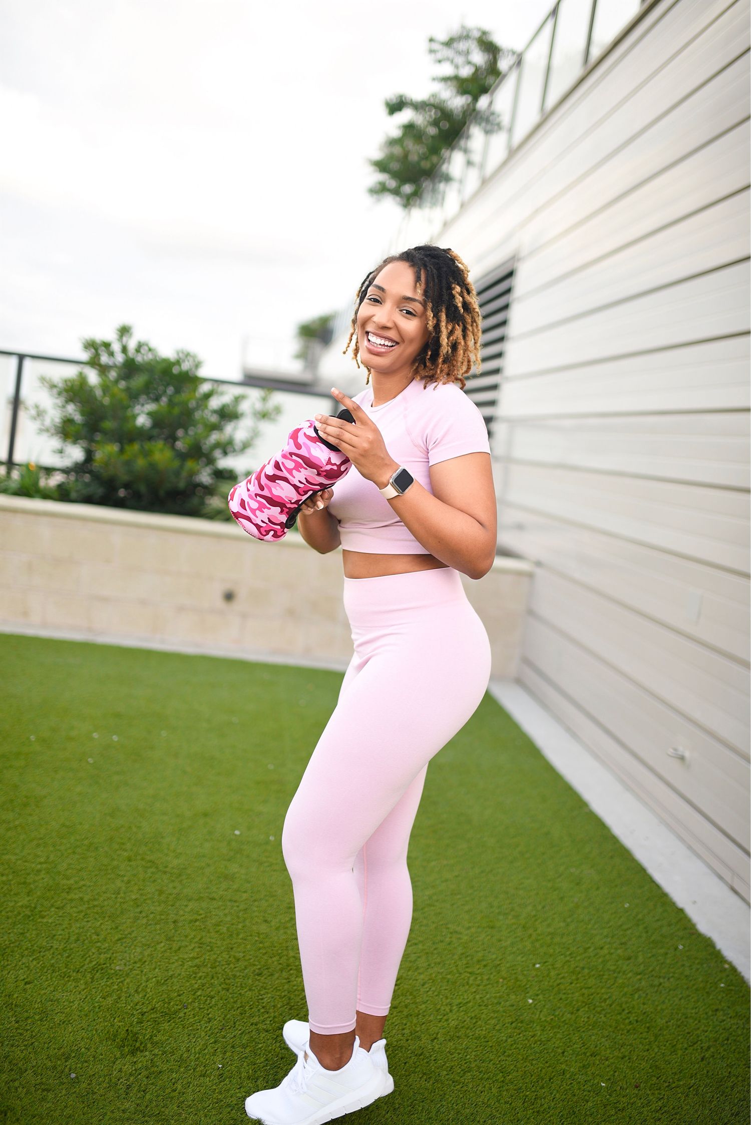 Woman in pink workout set holding a foam roller while smiling on a rooftop turf.