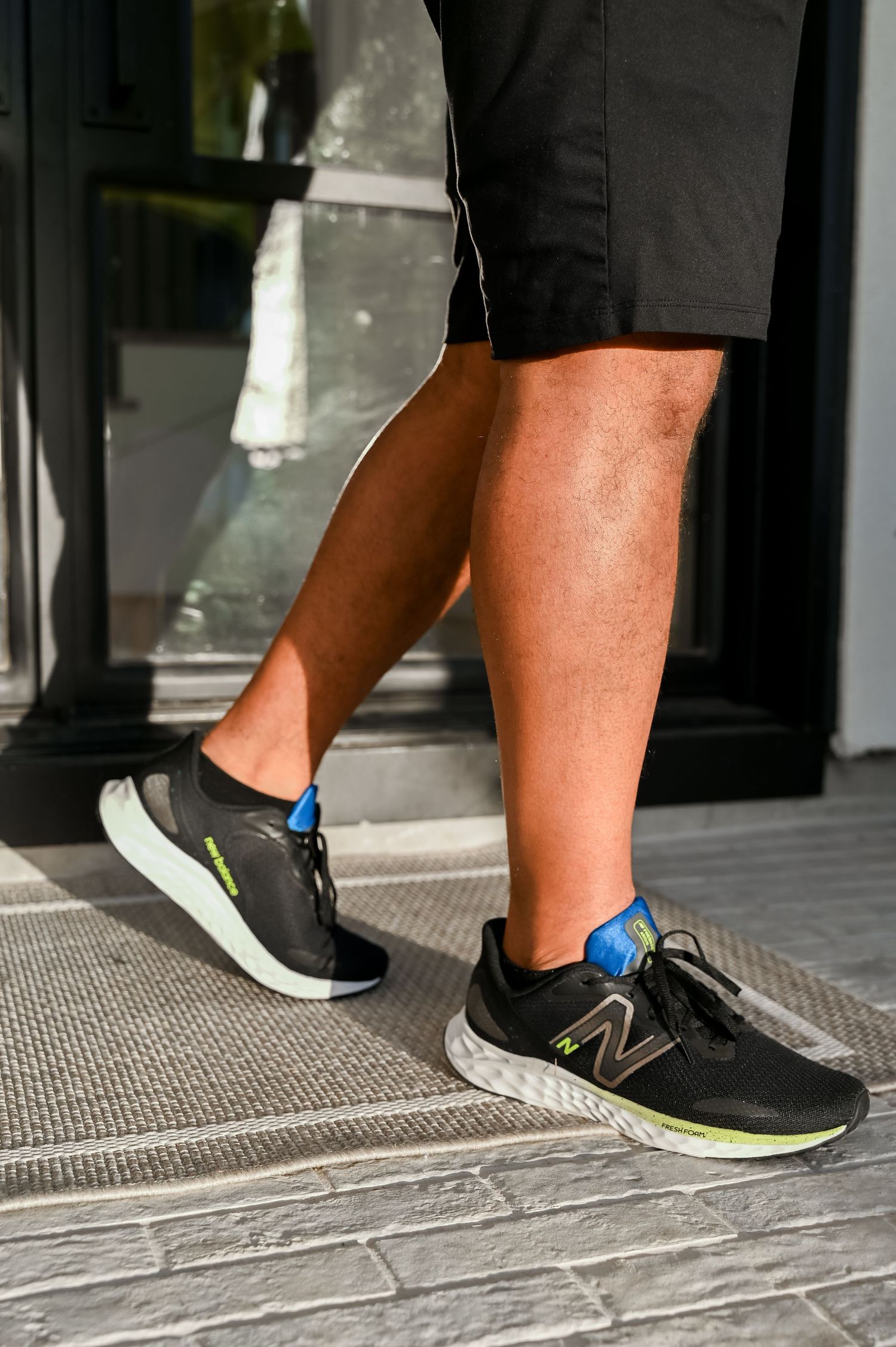 Man wearing black New Balance running shoes with neon green soles and blue accents on gray steps.