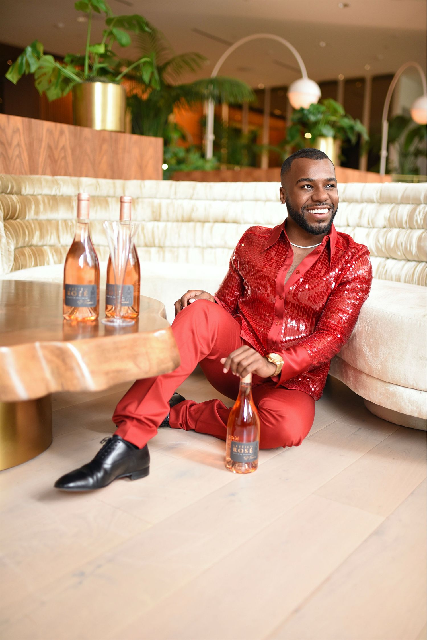Smiling man in red sequin outfit sitting on floor with rose wine bottles.