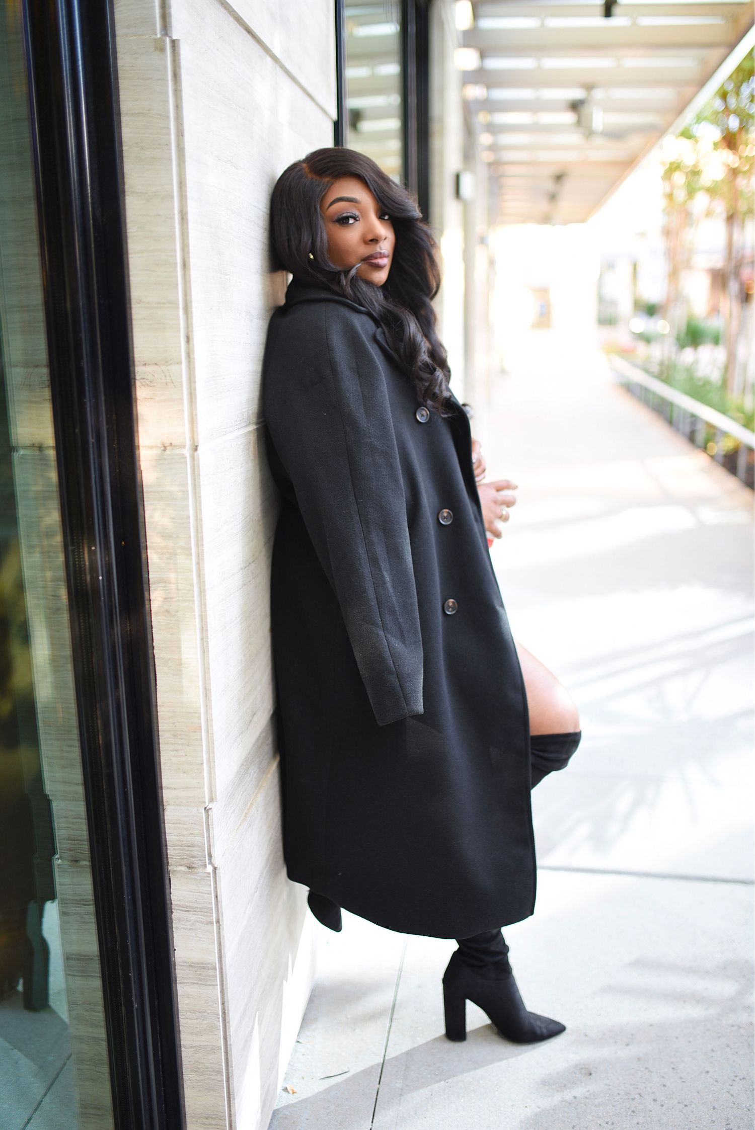 Stylish woman in long black coat and heeled boots leaning against marble wall outdoors.