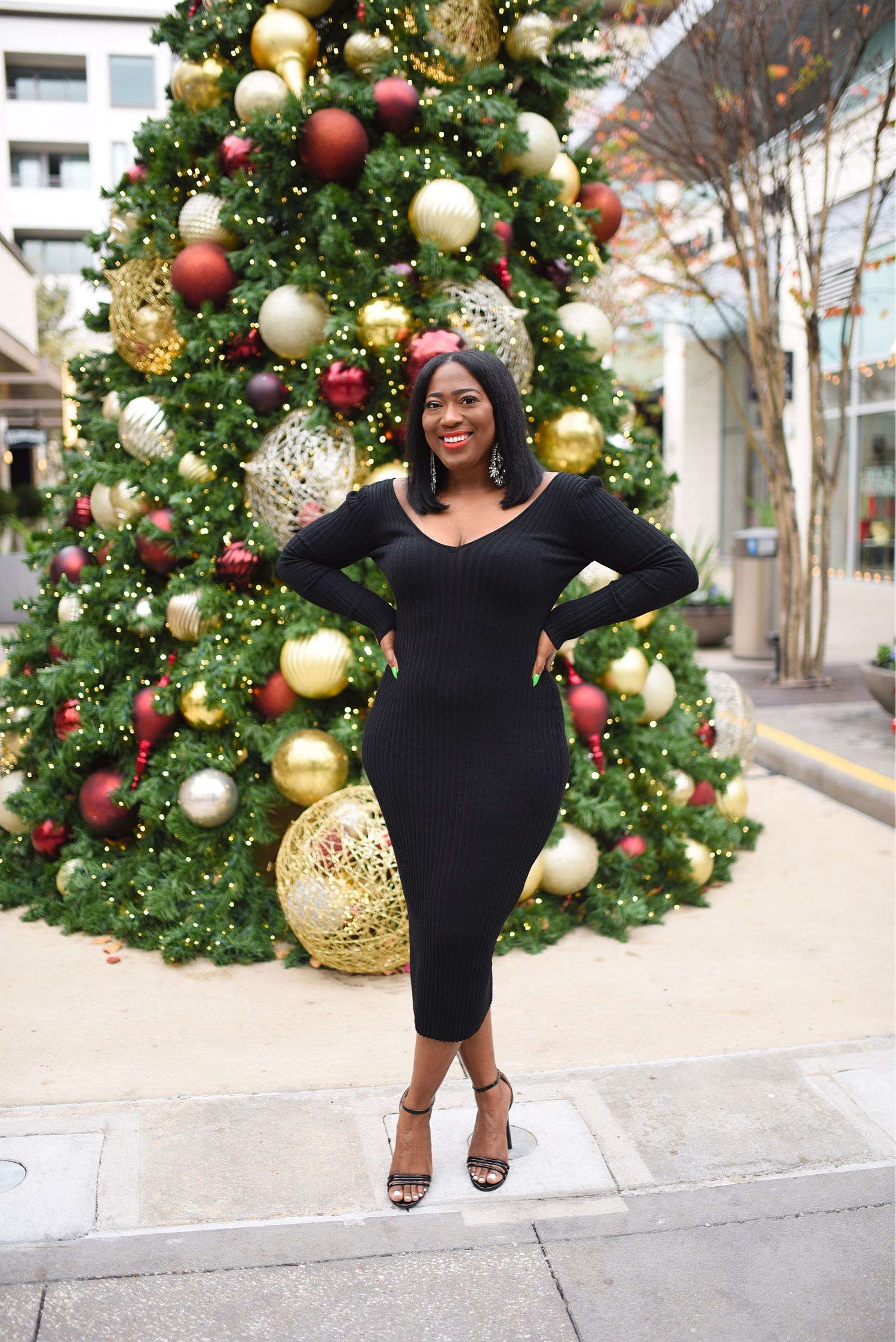 Woman in black midi dress posing in front of a decorated Christmas tree outdoors.