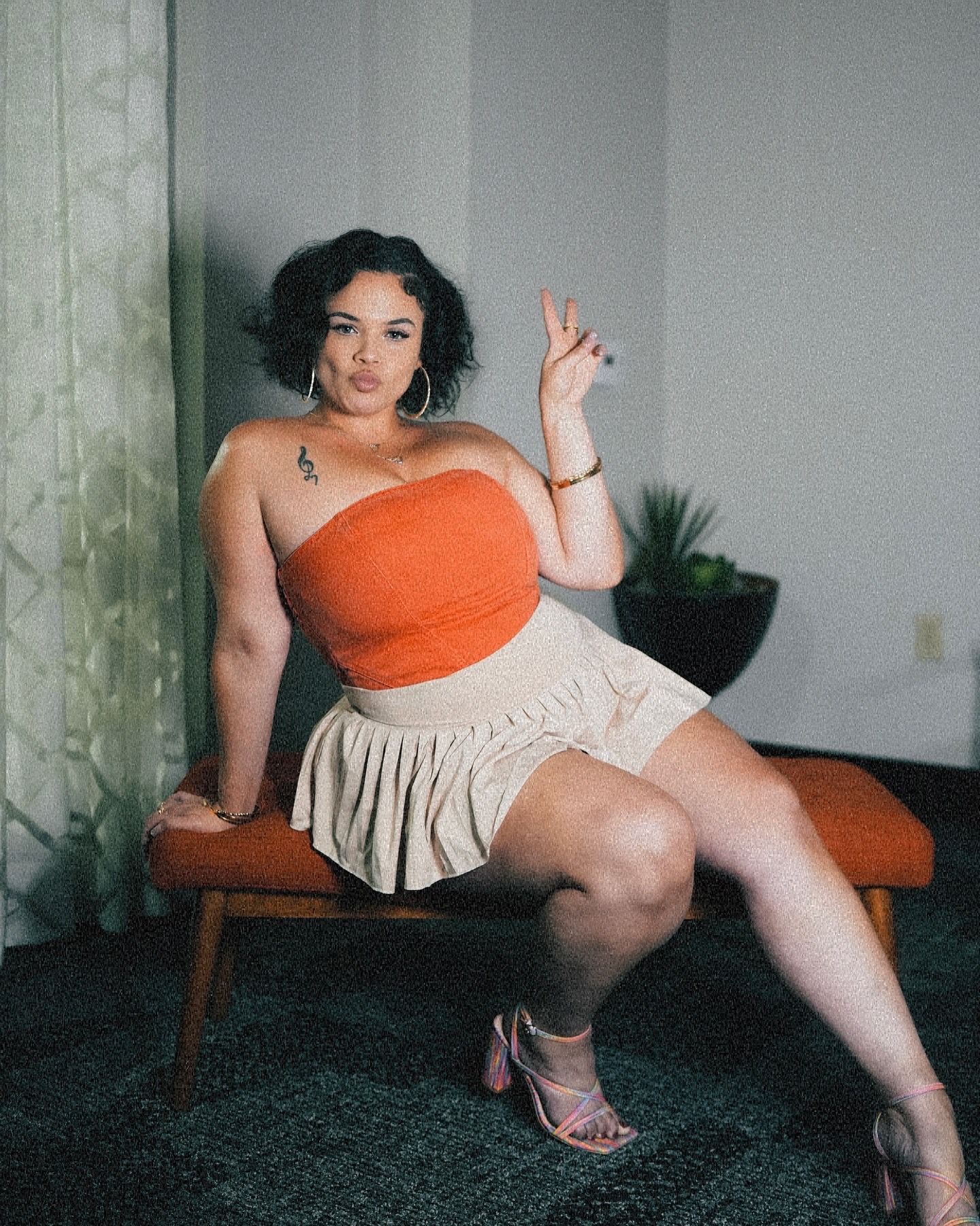Woman in orange strapless tube top and cream pleated mini skirt sits on a bench giving a peace sign.