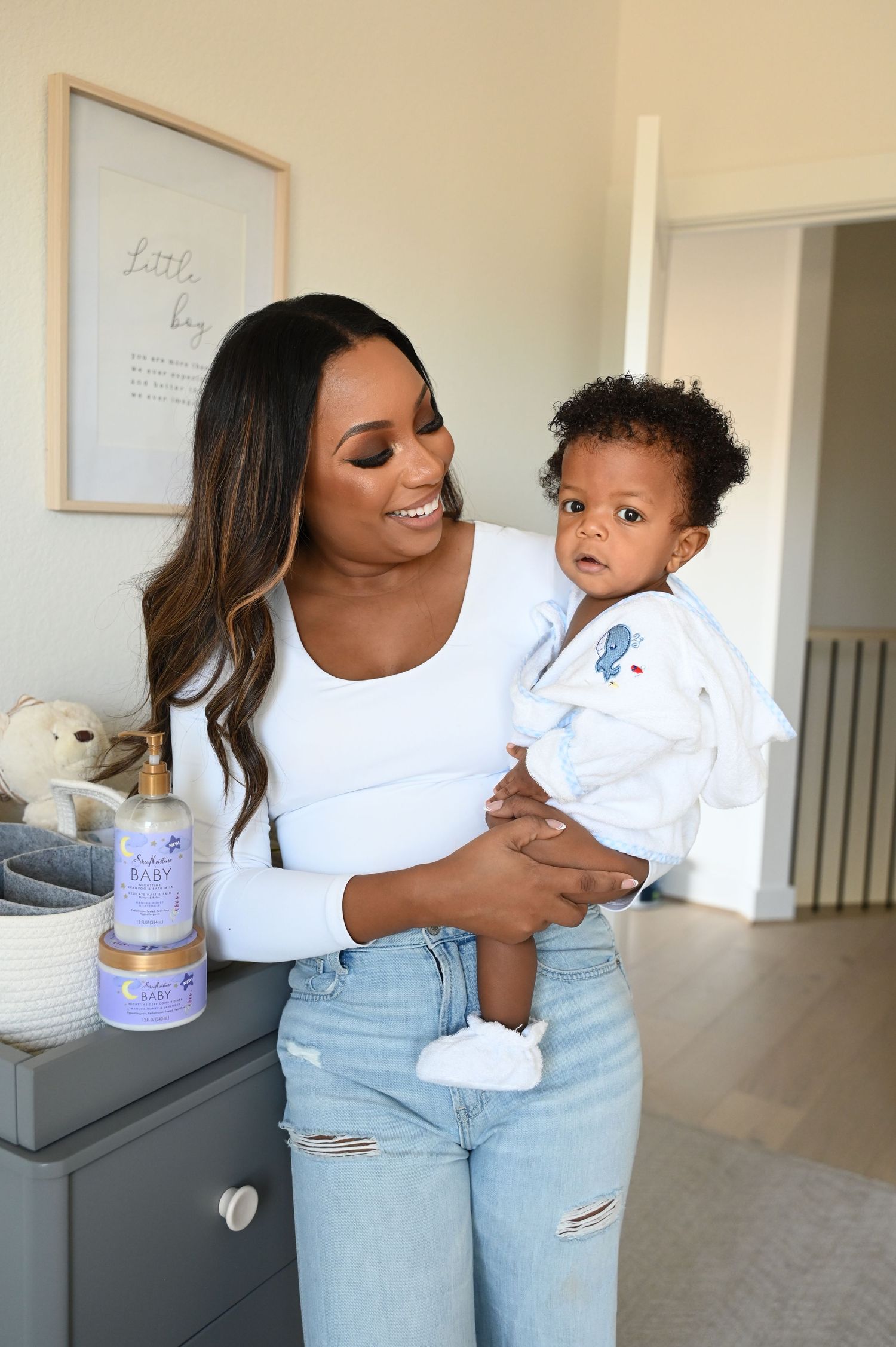 Smiling woman in white top holding a curly-haired baby near a dresser with baby lotion.