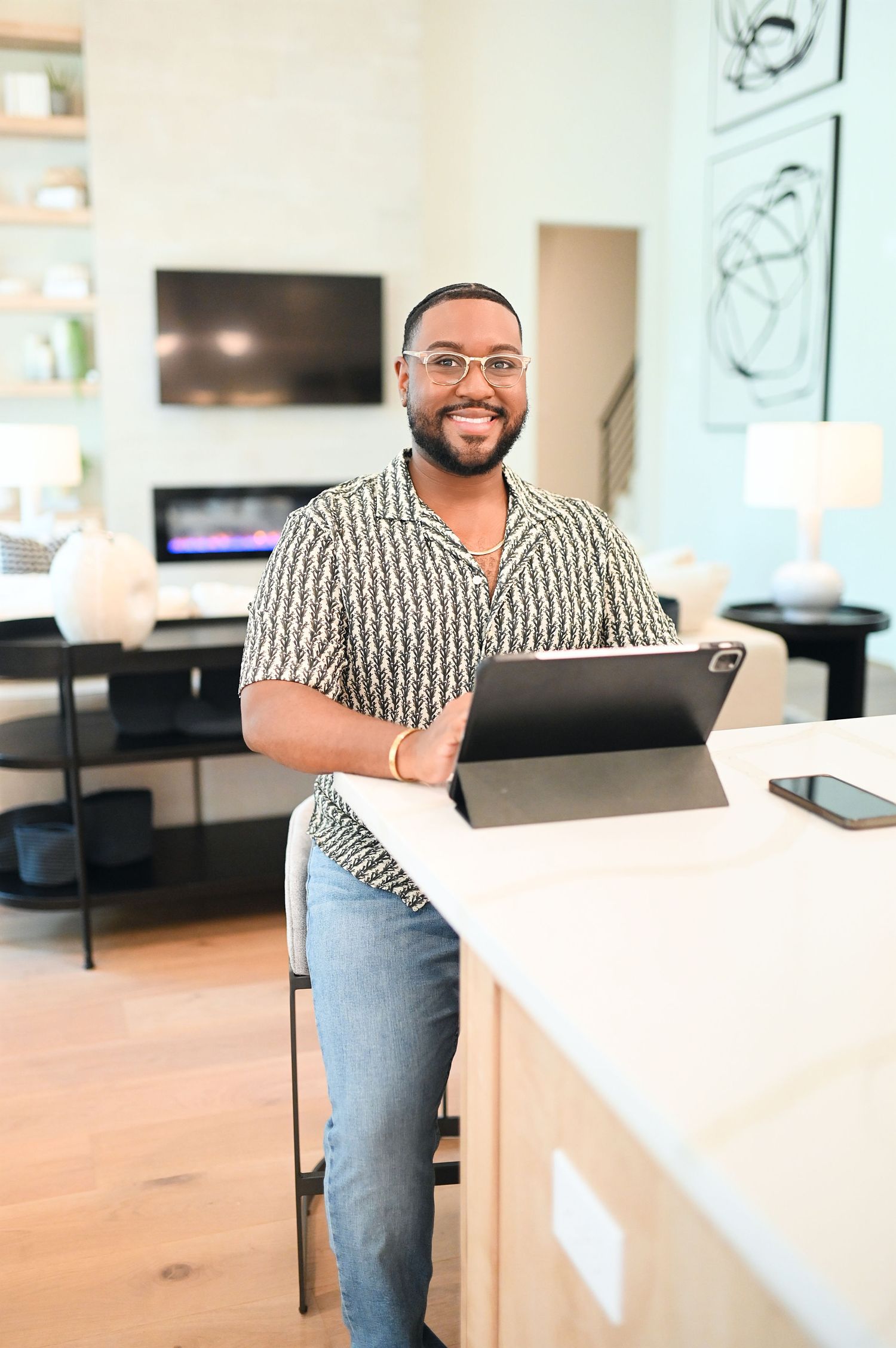 Smiling Black man in patterned shirt holds tablet while leaning on counter in modern home.