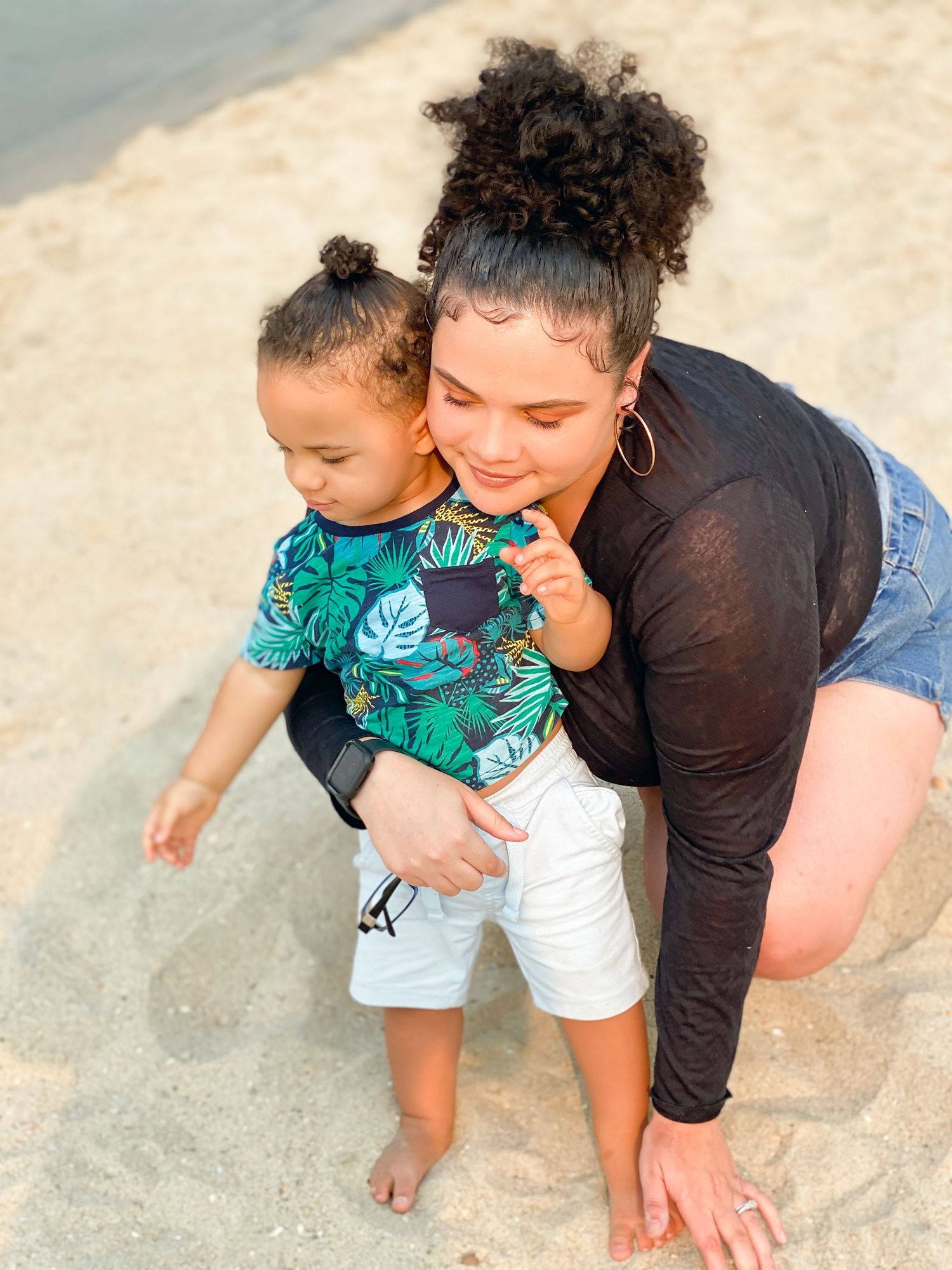 Young mother crouches on sandy beach with toddler in tropical shirt.
