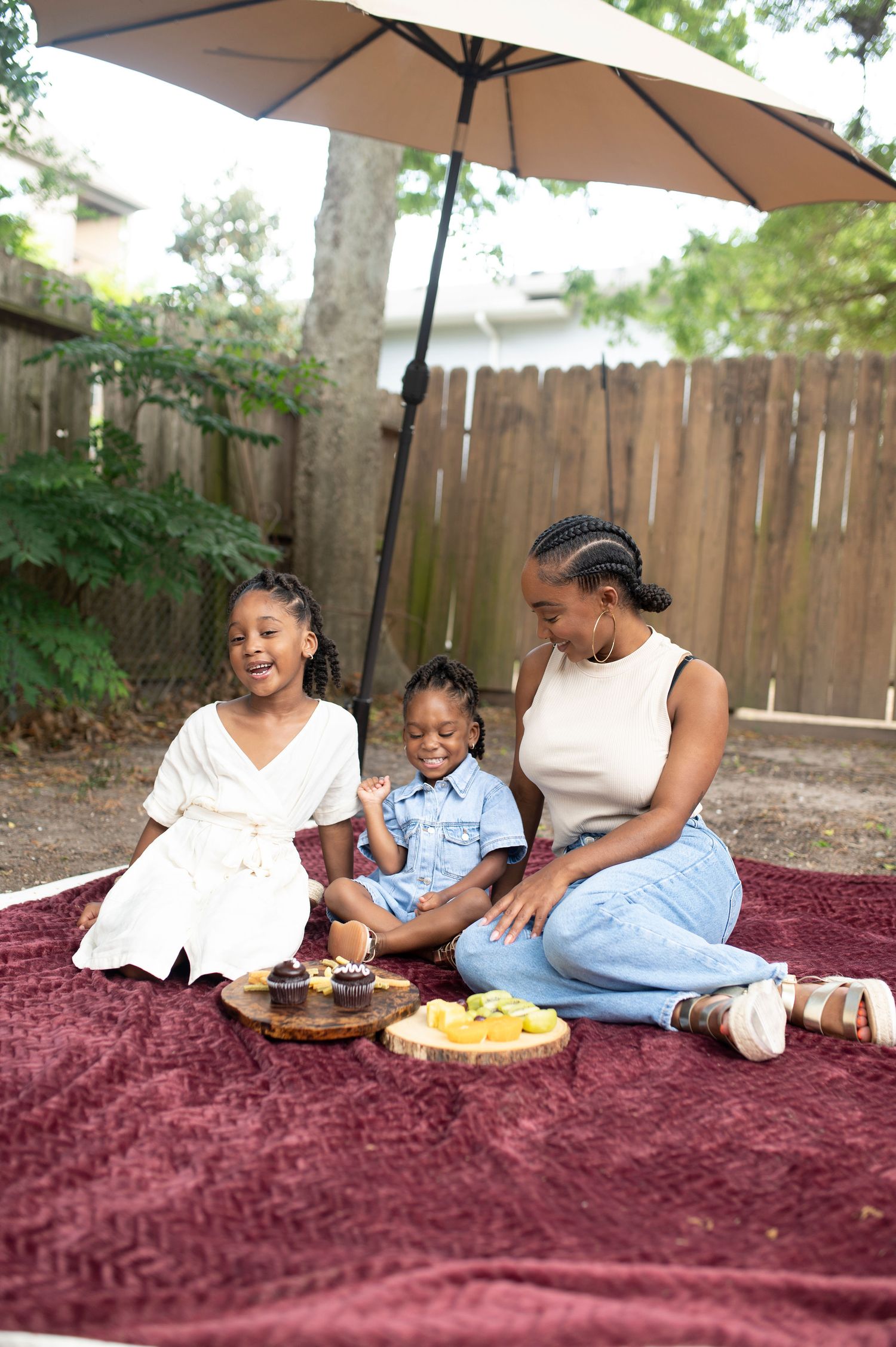 Happy family of mother and two children laughing together at an outdoor picnic with snacks under a patio umbrella.