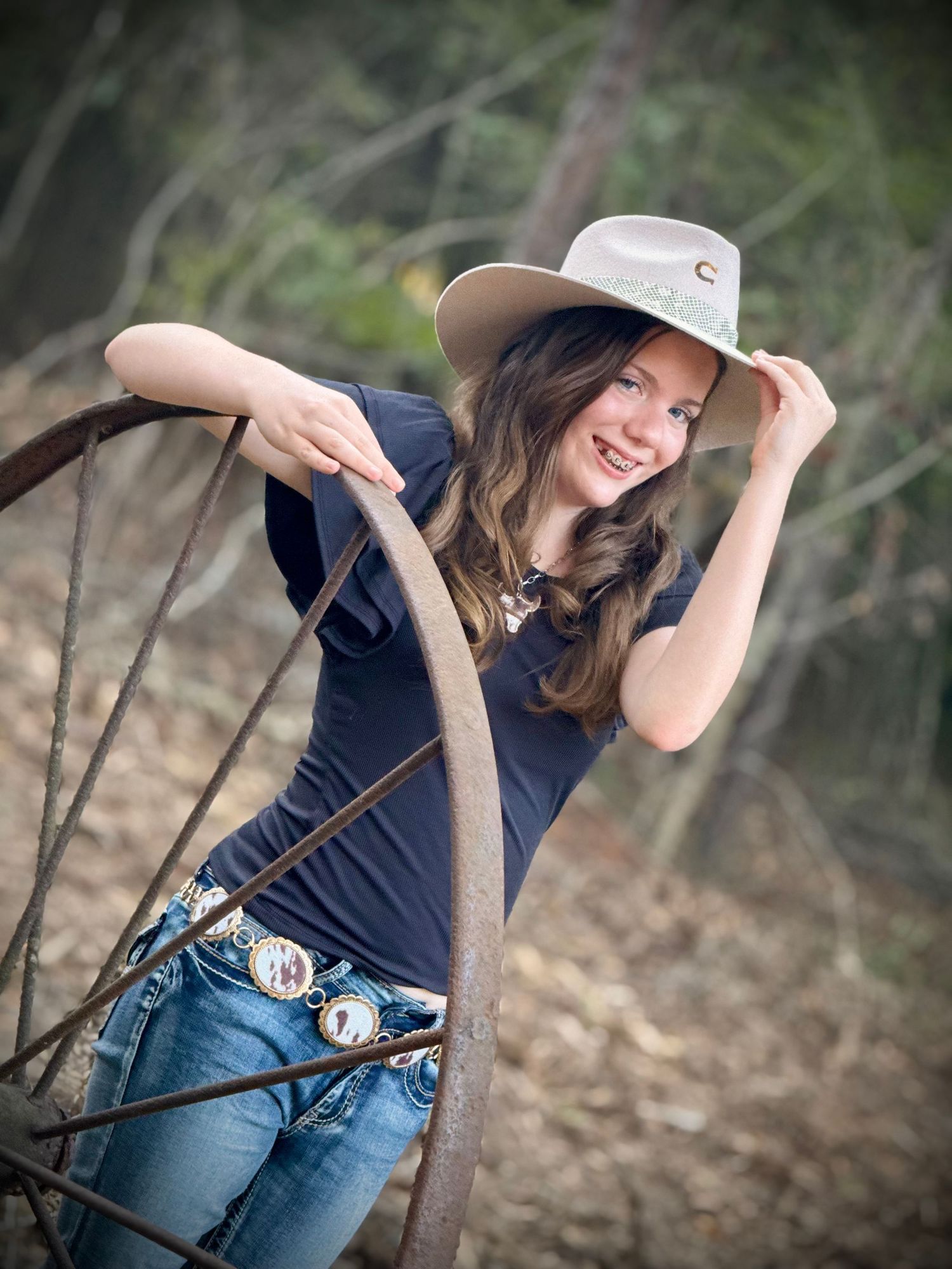 Teenager in navy shirt and jeans poses playfully outdoors with hat against natural wooded backdrop.
