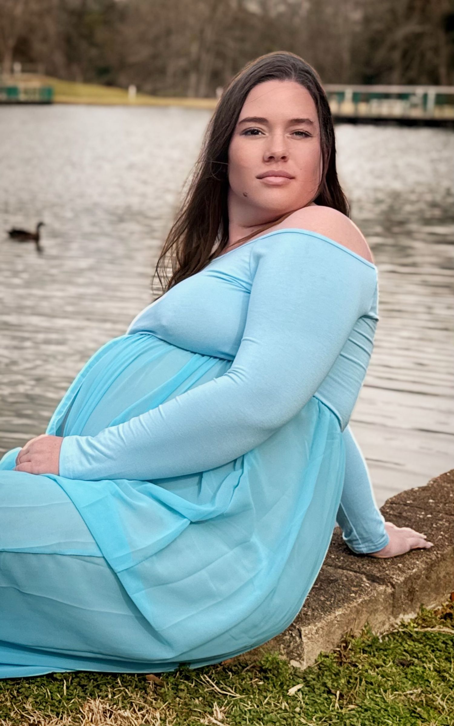 A mom to be in a light blue dress poses while laying on grass near a pond with ducks in the background.