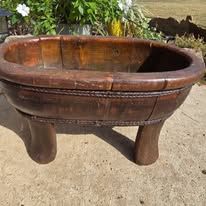 Vintage wooden oval bowl with carved details and three short legs stands on concrete surface in garden setting.