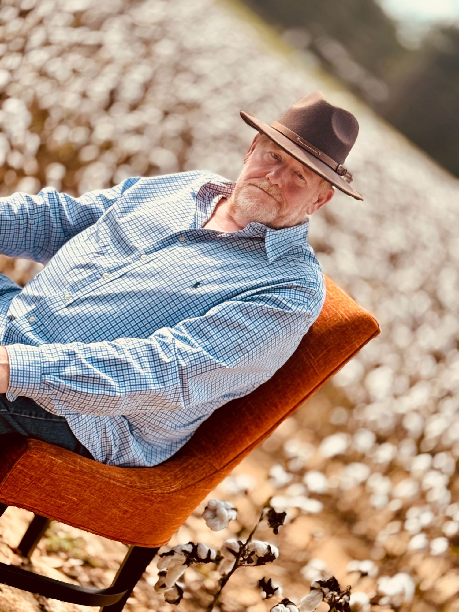Gentleman in a blue checkered shirt and brown hat relaxing on a fall colored chair in a sunny outdoor setting, inside a cotton field.