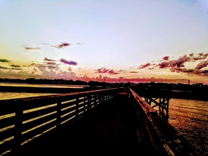 A wooden fence stretches along a rural landscape and water during sunset with pink and orange clouds in the sky.