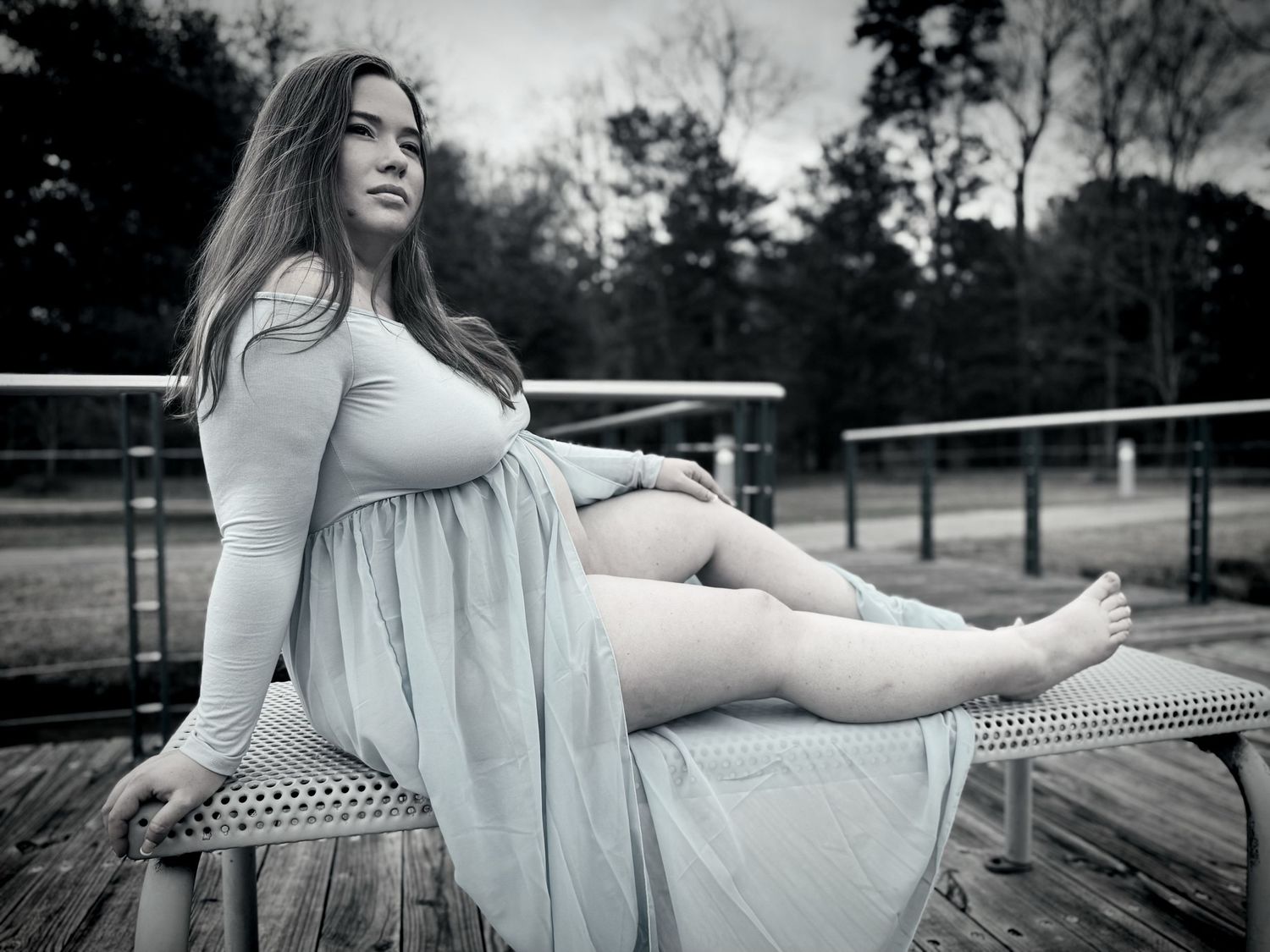 A woman in a light blue dress poses on a wooden deck during an outdoor surprise maternity photo session at sunset.