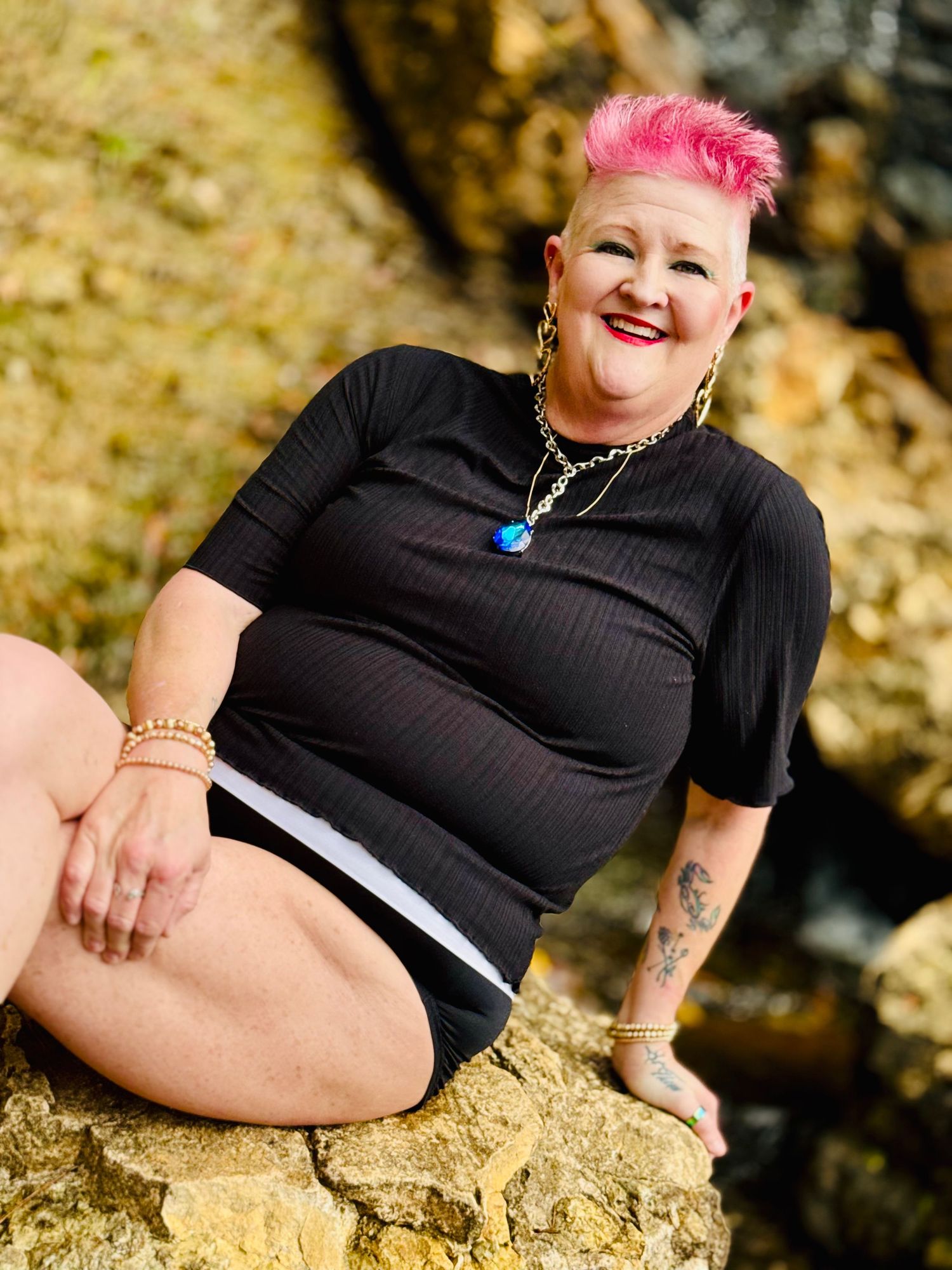 Amazing woman with pink hair wearing black clothing poses casually on rocky terrain in natural outdoor lighting.