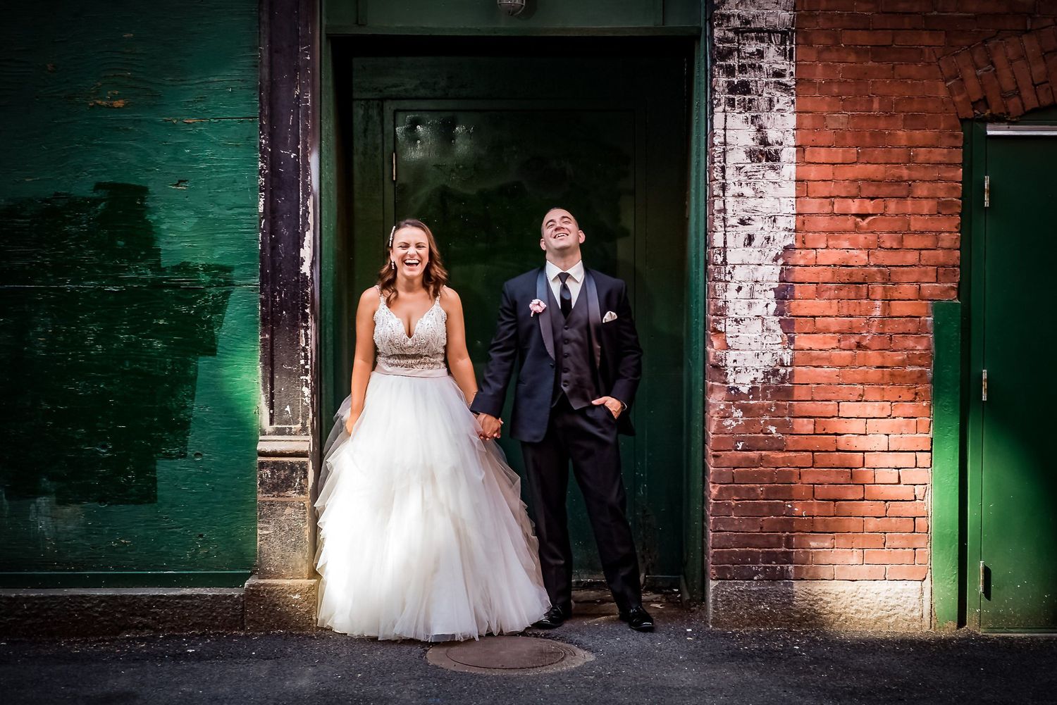 A bride and groom pose against a rustic brick wall with green doors. Taken by Rhode island wedding photographer