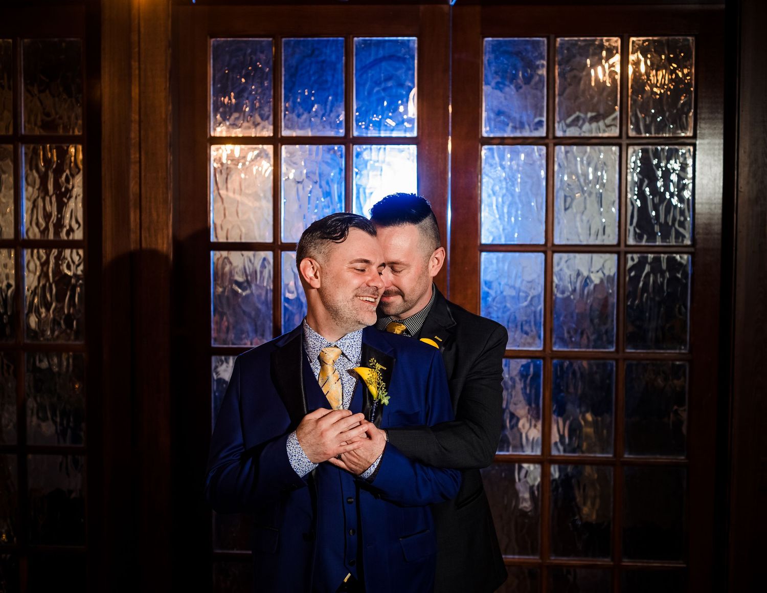Two people in navy blue suits share an intimate moment in front of a frosted glass window at night.