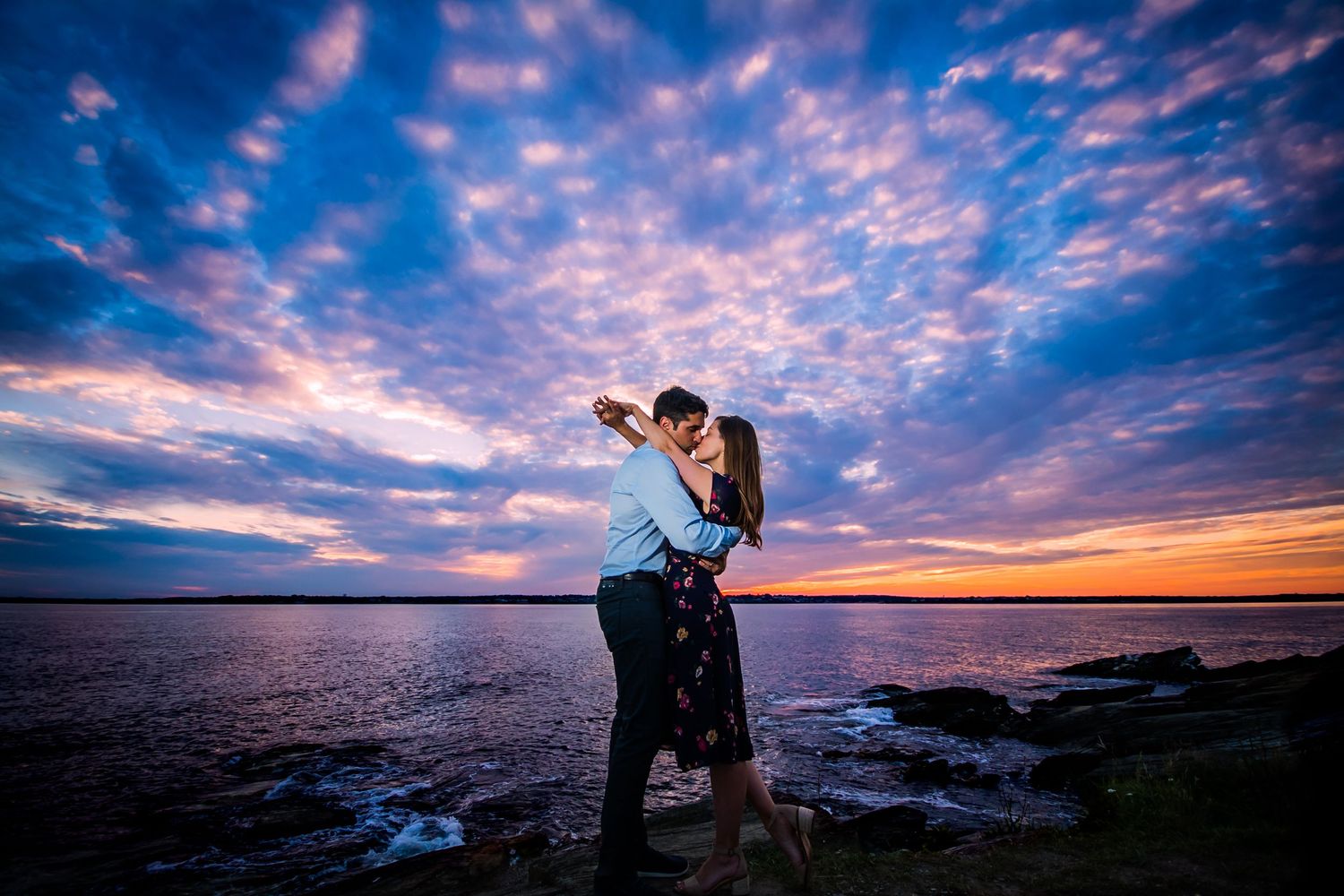 Dramatic sunset sky with pink and purple clouds over rocky coastline creates romantic backdrop, photographed by a Rhode Island photographer