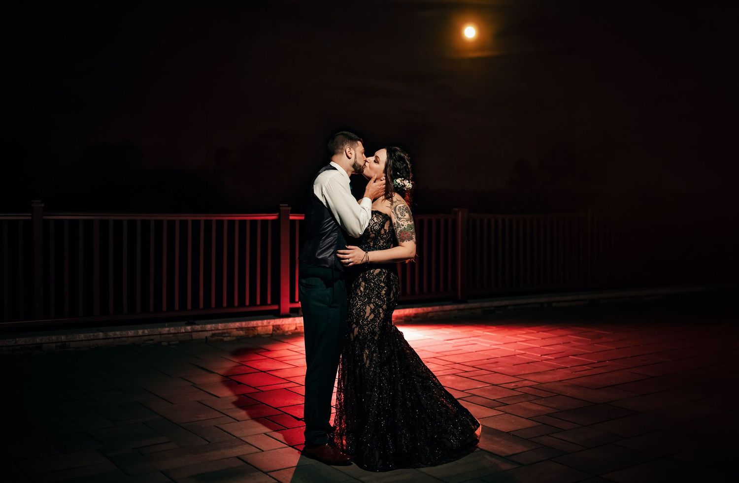 Couple sharing romantic kiss under full moon at night, woman in black lace gown, dramatic red lighting on rooftop.