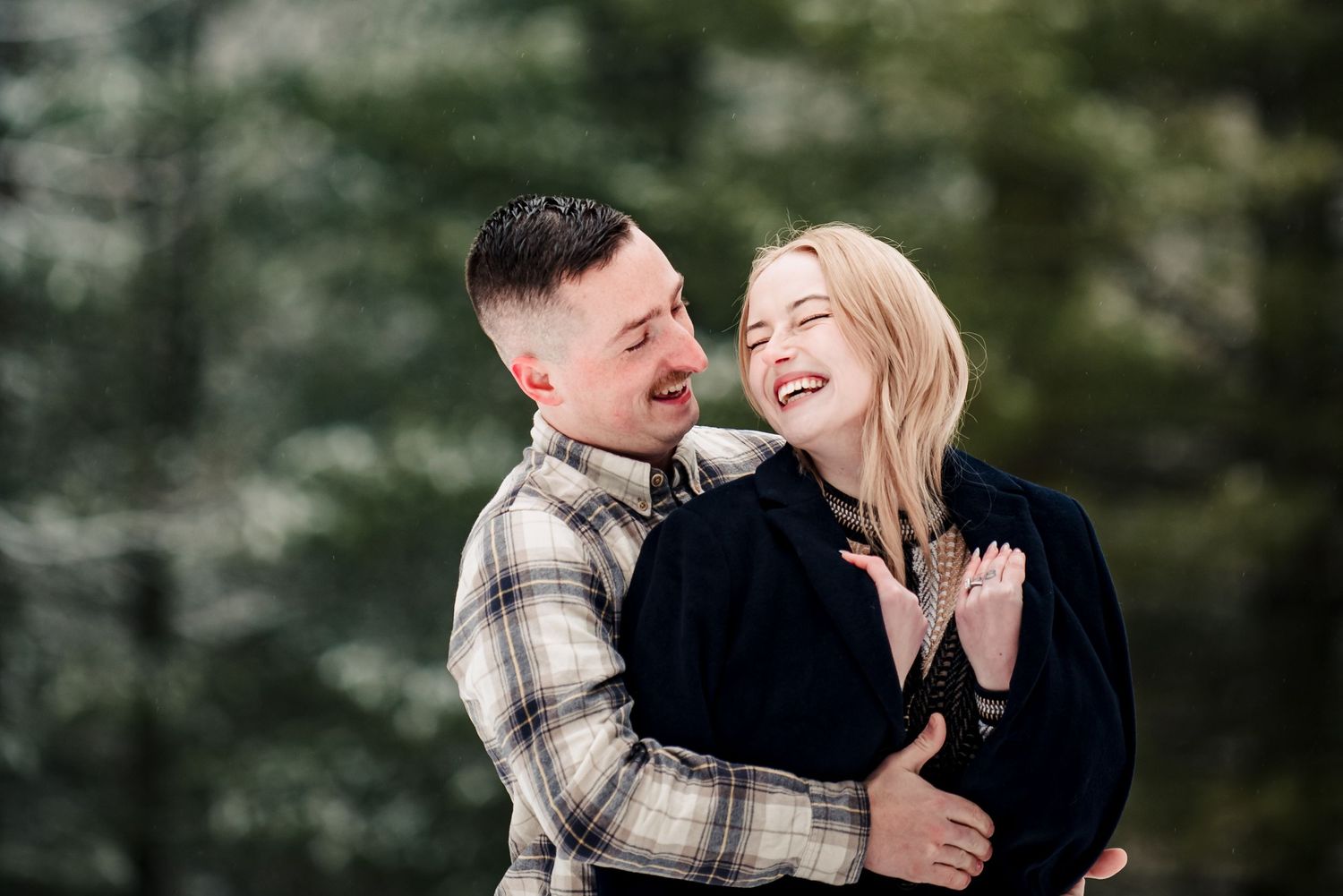 A laughing couple embraces outdoors in front of evergreen trees, wearing casual plaid and dark coats.