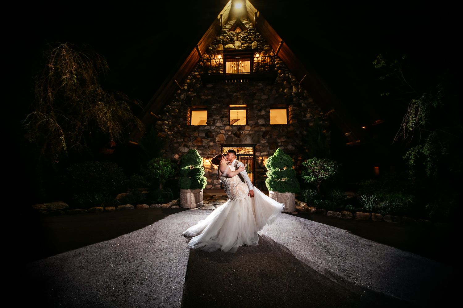 Bride and groom share romantic kiss outside stone chapel venue at night, wedding dress flowing dramatically.