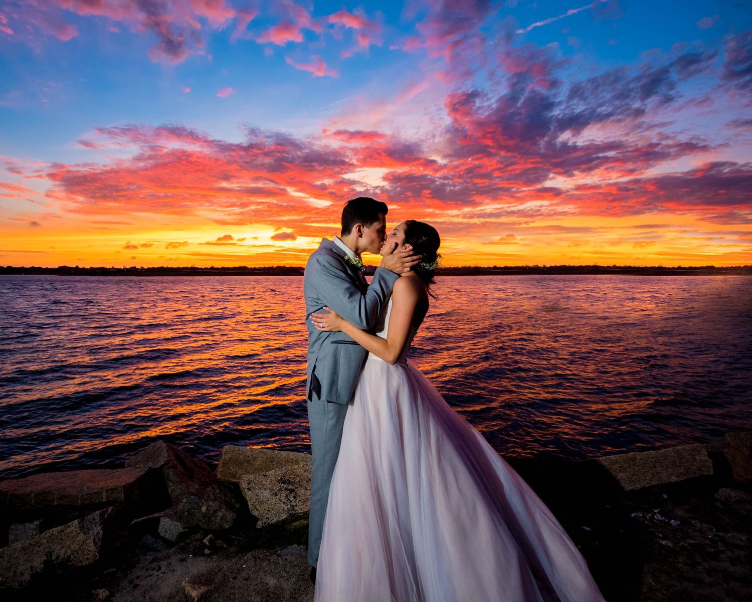 Dramatic sunset wedding photos along waterfront with vibrant pink and orange sky, photographed by a Rhode Island wedding photographer