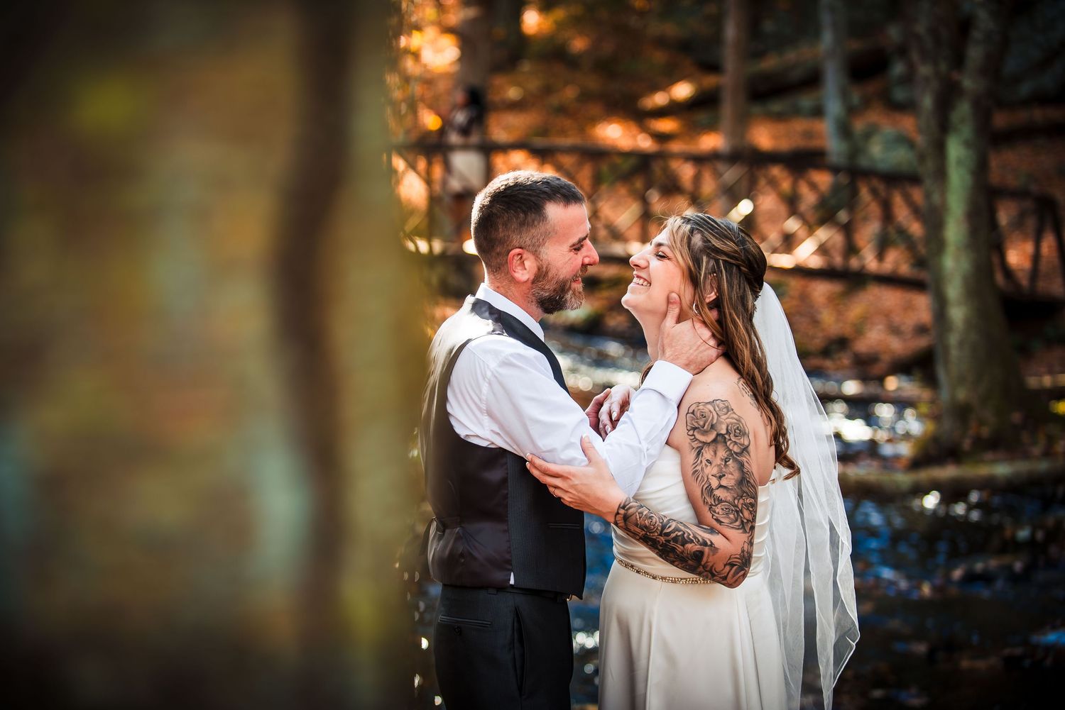 A tattooed bride and groom share a tender moment outdoors among autumn trees at their wedding.