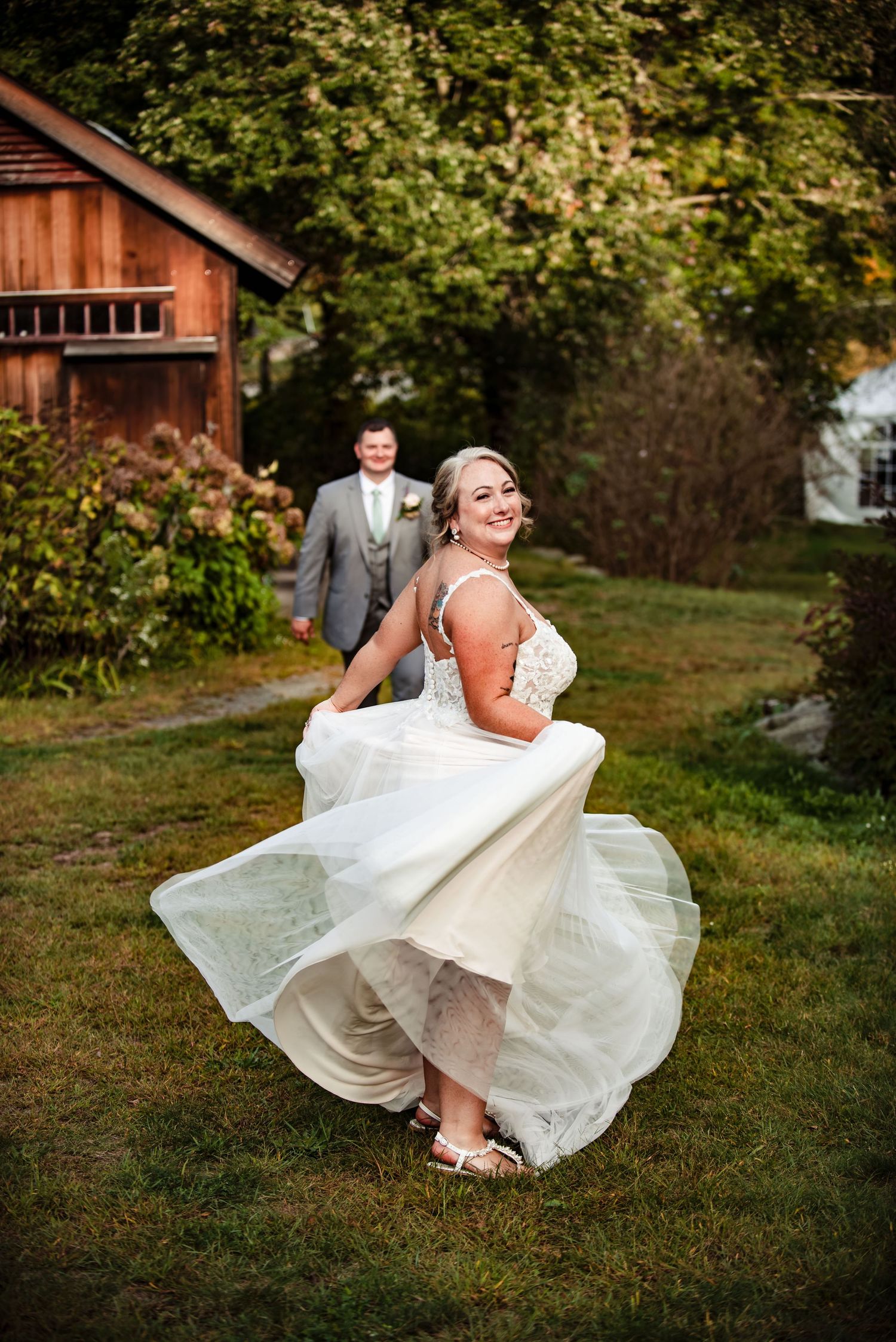 A bride twirls her white wedding dress on a grassy lawn with a rustic barn in the background. Taken by Rhode Island wedding Photographer