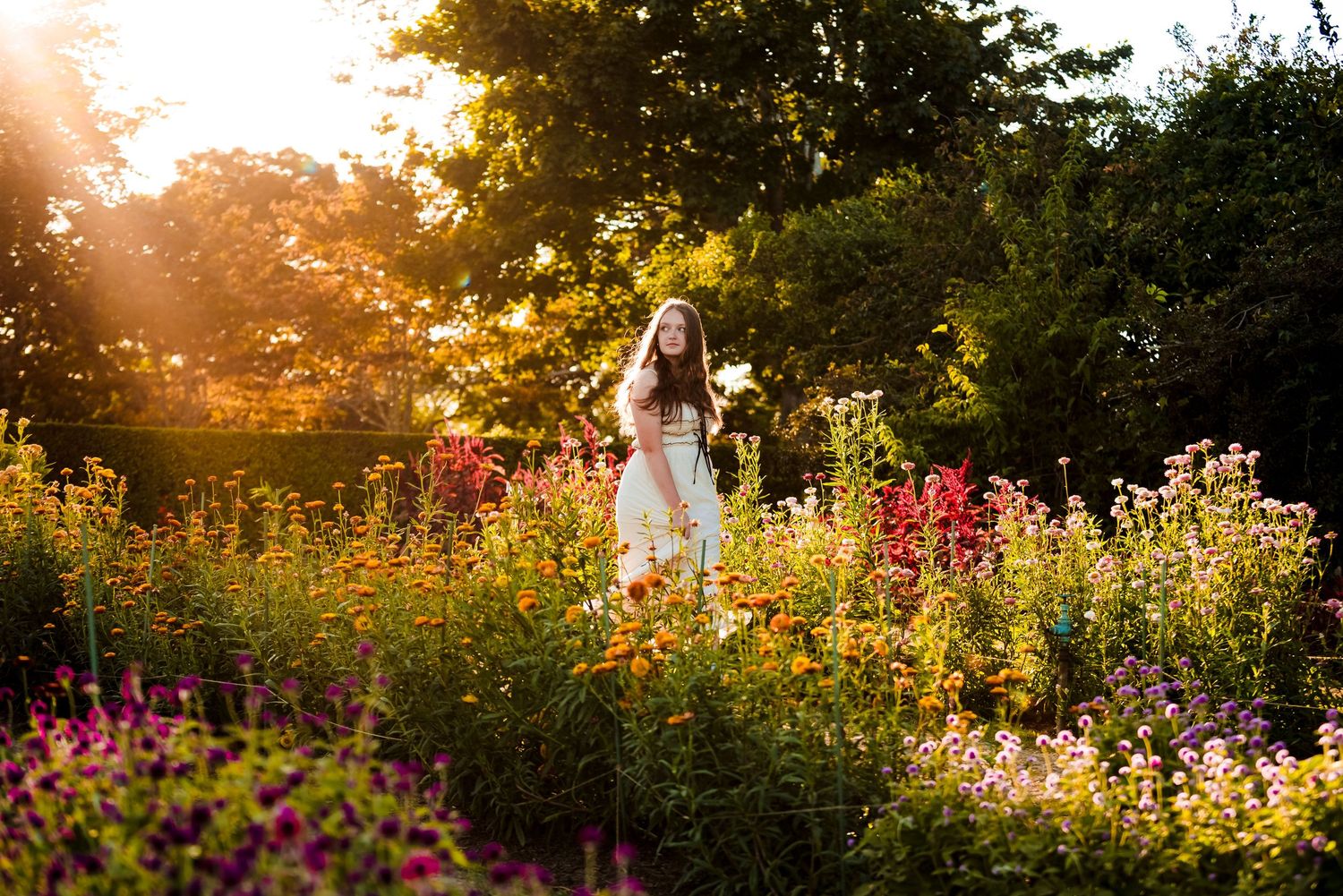 A figure in a white dress stands among vibrant wildflowers in a garden during golden sunset light.