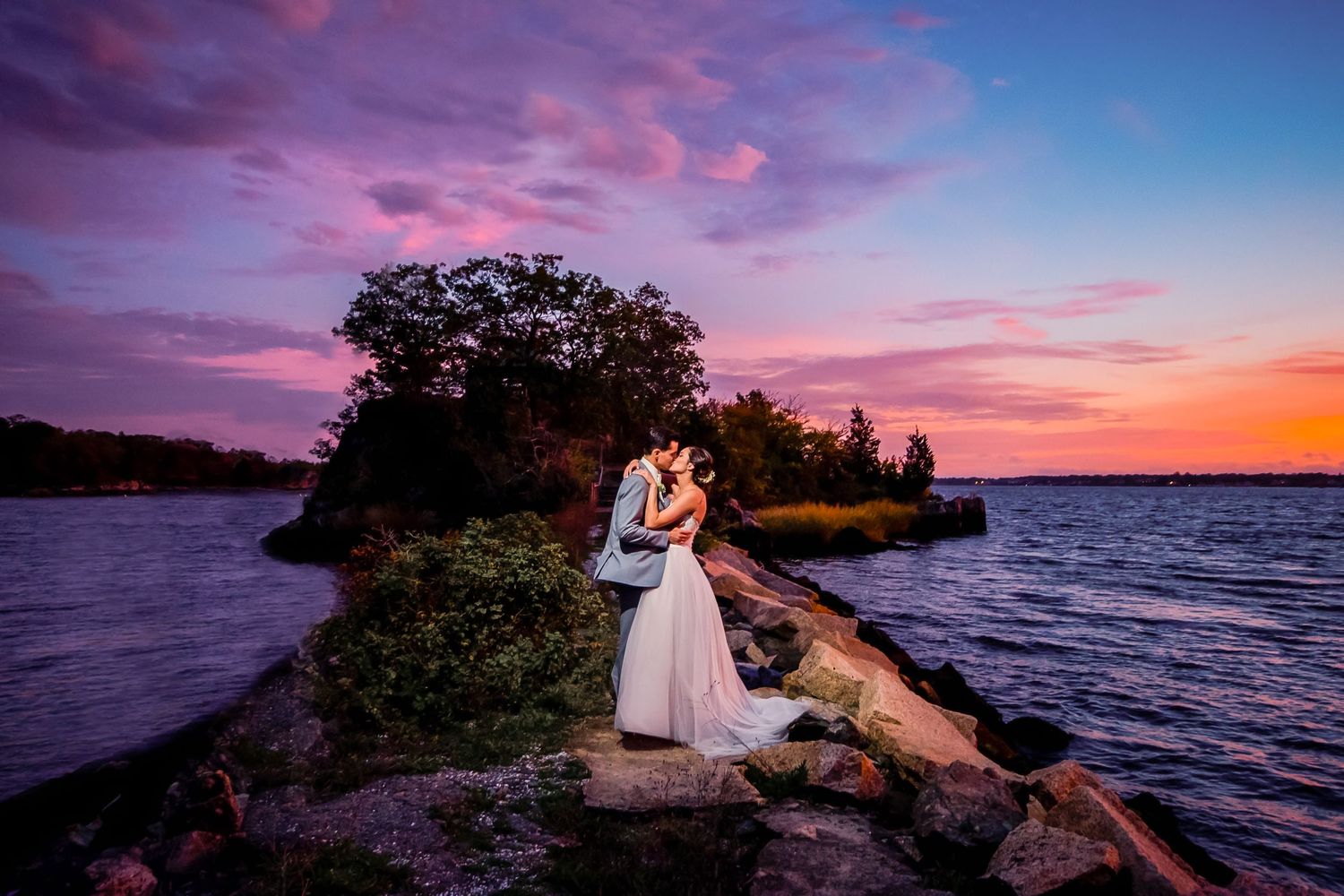 Couple in wedding attire embraces against a vibrant pink and purple sunset sky over a serene waterfront landscape.