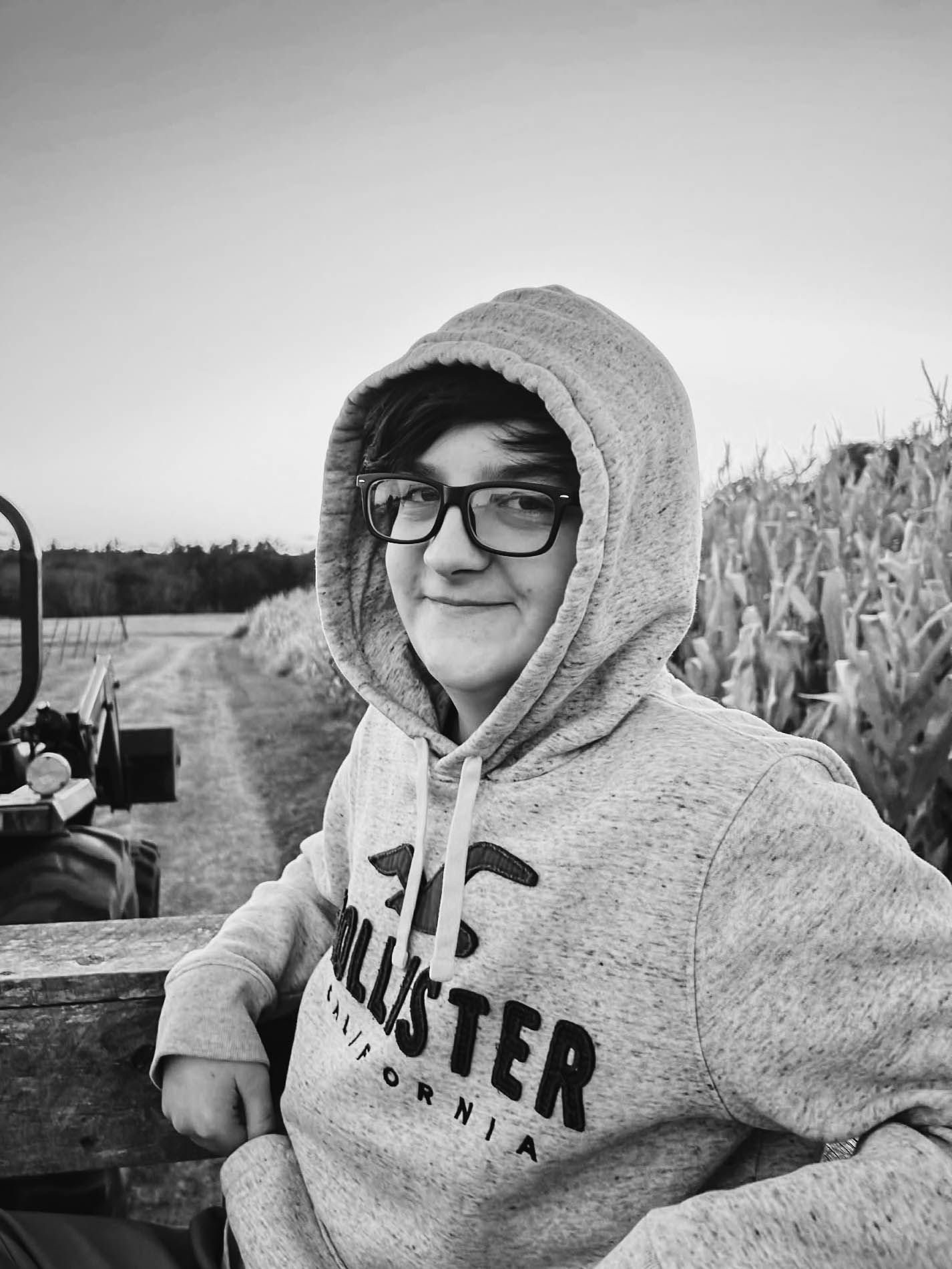 Person in gray hoodie and glasses posing outdoors near cornfield.