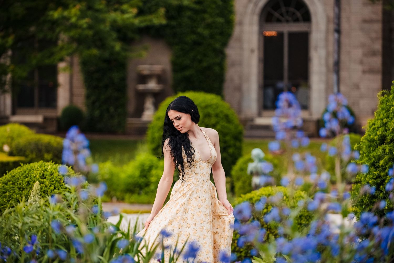 A woman in a cream dress stands in a garden with blooming blue delphiniums and a stone building in the background.