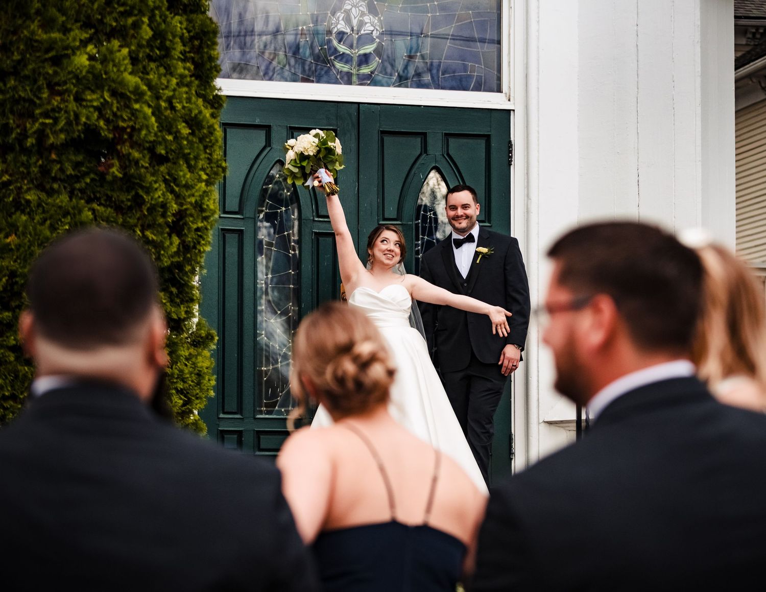Joyful bride raising bouquet with groom at green church doors, greeted by wedding guests outside.